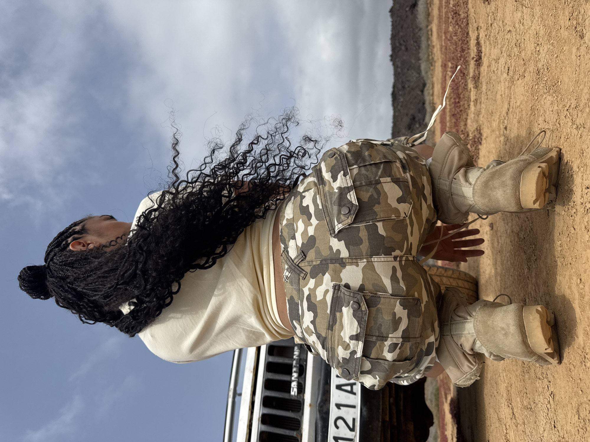A medium, full shot presents a person kneeling near a vehicle on a tan, dirt-like ground, with a backdrop suggesting an outdoor location in Tinajo, Spain, characterized by its arid landscape and volcanic terrain. The ground transitions to a ridge of multicolored earth. The subject, positioned with their back towards the viewer, is wearing camouflage pants, a cream-colored top, and tan boots with laces. Their dark, long, curly hair is styled in braids. The partially visible black vehicle behind them has a white license plate reading "121A." Above, the sky is partially overcast with patches of blue.