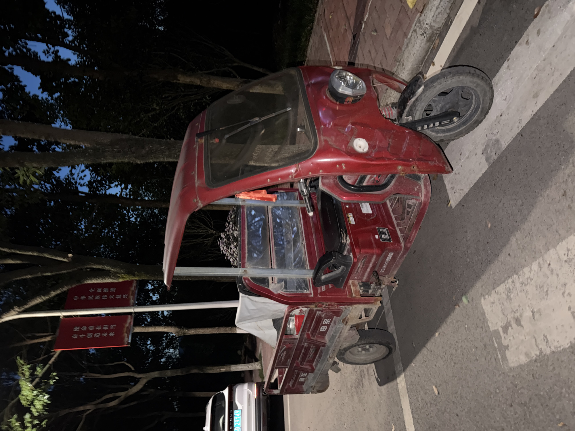 An outdoor, eye-level, medium shot captures a red, three-wheeled motorized vehicle parked on the side of a paved road at dusk. The vehicle is facing to the right, with its front wheel visible. The cabin of the vehicle is open on the driver's side, revealing a black seat and steering wheel. The rear of the vehicle is a cargo bed covered with a white tarp.
In the background, several trees with dark foliage line the road, and a lamppost with two red banners is visible. The banners contain Chinese characters. The sky is dark blue, indicating evening. To the left of the three-wheeled vehicle, a white SUV with a blue license plate is parked. The road surface is asphalt with white markings. The scene appears to be in Jiangjin, China, suggested by the presence of the banners, which are often used for public information and propaganda in Chinese cities. No people are visible in the image. The lighting suggests artificial illumination from streetlights or the vehicle's headlights, though the headlights appear to be off. The overall atmosphere is quiet and still.