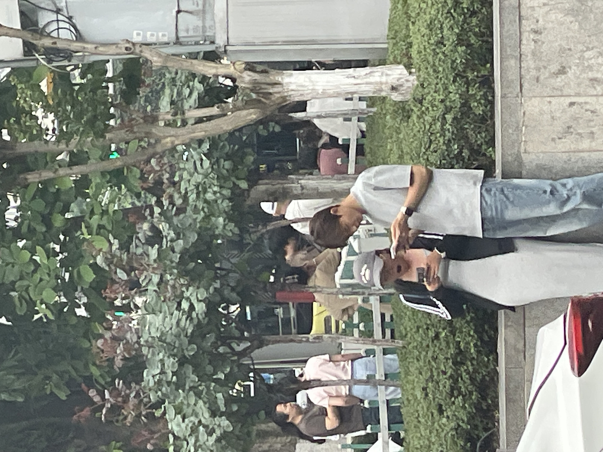 The image shows a group of people standing or sitting along a sidewalk in what appears to be an urban setting. Trees and greenery are visible, suggesting a park or landscaped area. Several individuals are seen casually dressed, engaging in conversation or using their phones. In the background, there is a building and parked cars, and some benches are present near the people. The location is suspected to be Yuzhong District, China.