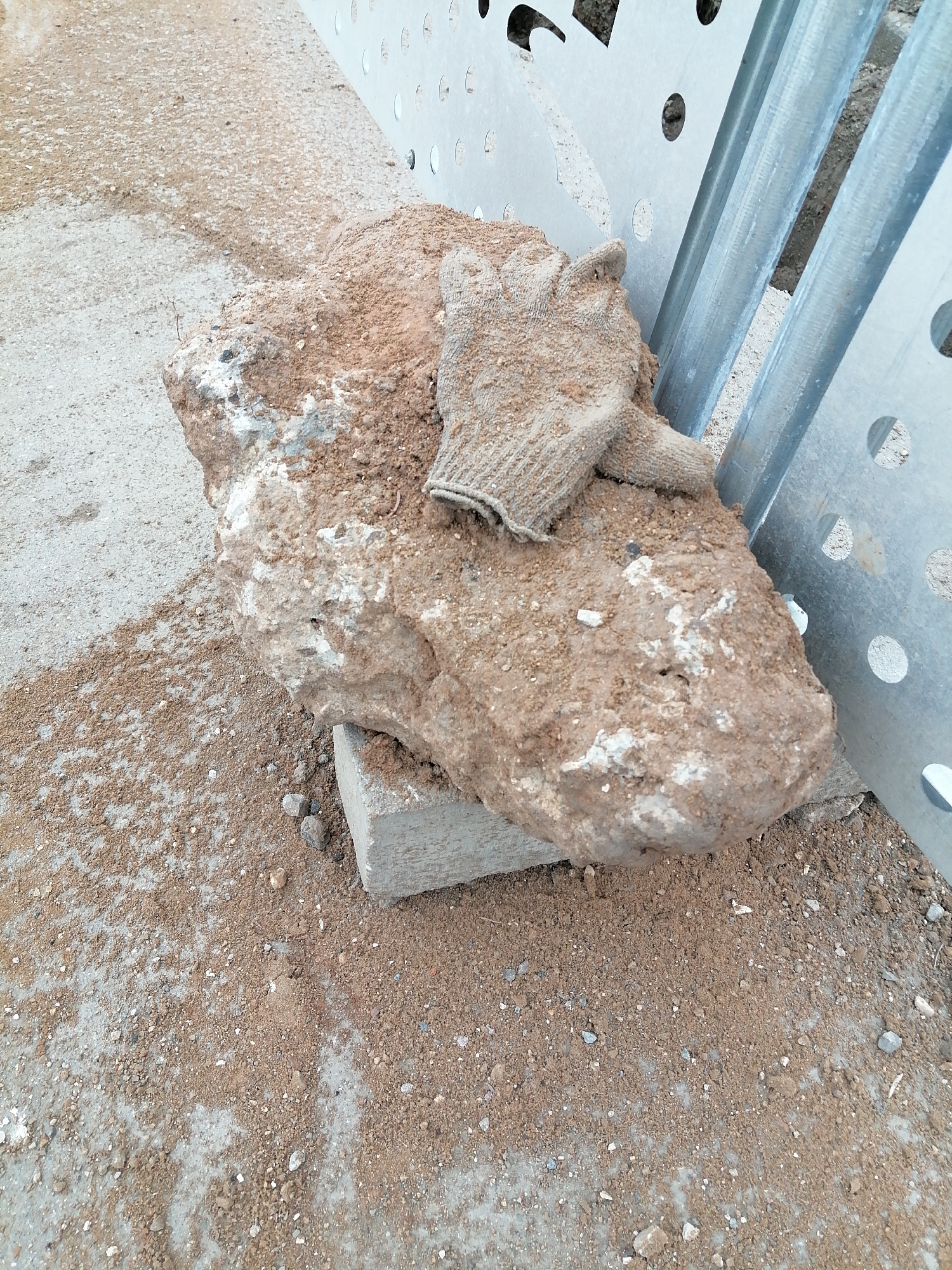 A large, irregular rock, coated in reddish-brown dirt, rests on a small, gray concrete block. A dirty, tan-colored work glove sits atop the rock. The rock and block are positioned on a concrete surface covered with loose dirt and small pebbles. To the right of the rock, a section of perforated metal framing is visible, constructed of steel. The setting is likely an outdoor construction or maintenance area. There is insufficient information to locate this image in the city of Moscow, Russia.