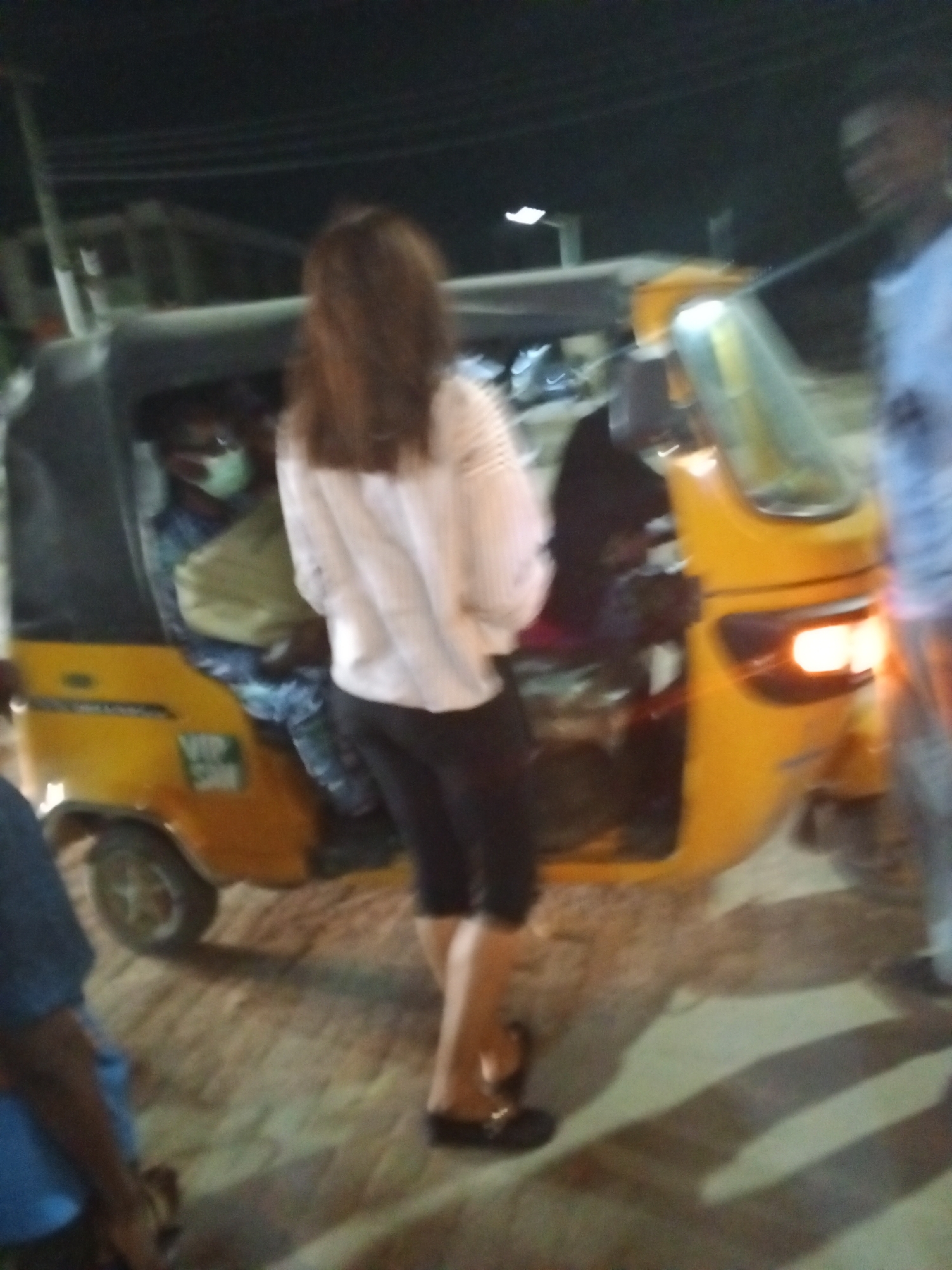 The image captures a nighttime scene, likely on a street or roadside in an urban or semi-urban area. The primary focus is a yellow auto-rickshaw, commonly known as a "keke Napep" in Nigeria, with a black canvas canopy. Its taillights or headlights are illuminated with an orange glow, indicating it is either operating or recently stopped.
In the foreground, a woman with long, brown hair stands with her back to the camera, positioned next to the auto-rickshaw. She is wearing a light-colored, long-sleeved shirt, black capri-style pants, and flat black sandals. Inside the auto-rickshaw, a passenger is visible, wearing a light blue face mask and what appears to be a light-colored garment over patterned clothing. Their posture suggests they are seated.
To the right of the auto-rickshaw, a blurry figure of a man in a light-colored shirt is partially visible. Another person's arm and shoulder, wearing a blue shirt, can be seen in the lower left corner of the frame. The activity appears to involve people gathered around the auto-rickshaw, possibly waiting to board, disembark, or simply interacting.
The ground is paved with what looks like interlocked bricks or concrete. The darkness of the sky, along with the illuminated lights of the vehicle and faint suggestions of distant streetlights or structures, confirms the scene takes place at night. Overhead wires are faintly visible against the dark backdrop. A small, white sticker on the side of the auto-rickshaw displays the text "VIP" along with some smaller, illegible markings. The overall image has a somewhat blurry quality, suggesting motion or low light conditions.