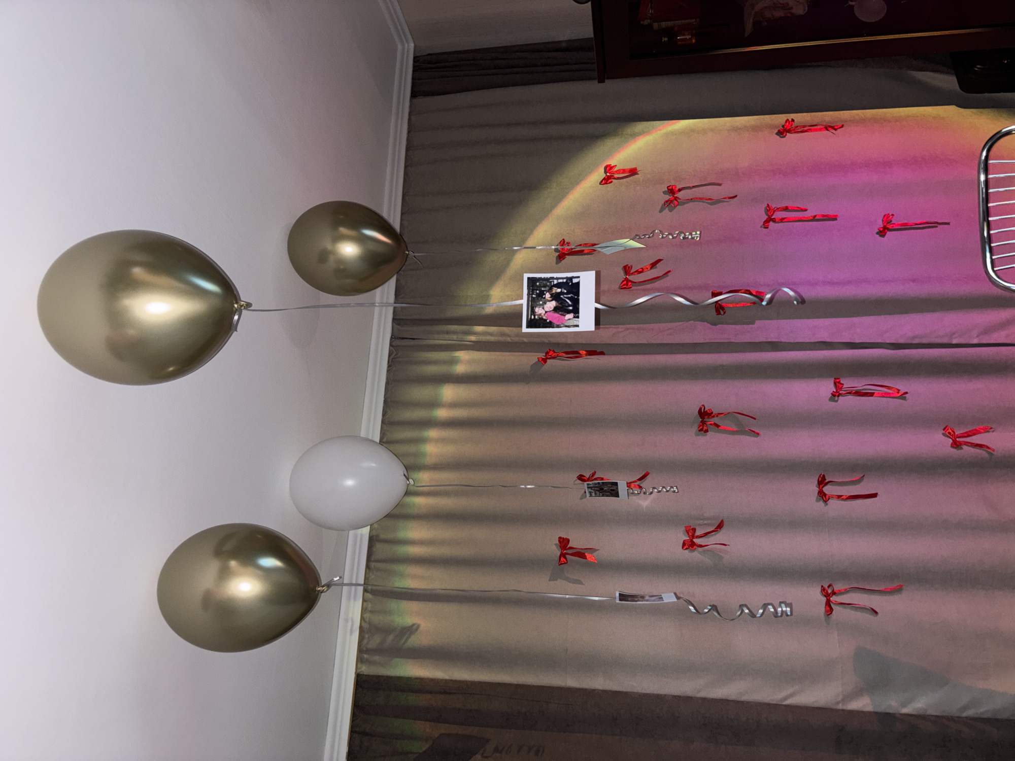 The image captures a room decorated with balloons and photographs, likely for a celebration. Two gold balloons and one white balloon are visible on the left, tethered by strings. The strings also hold up small square photographs. The background consists of draped fabric or curtains, decorated with small red bows, and illuminated with a gradient of pink and yellow light. A dark wooden cabinet or shelf is partially visible at the top. The location is not discernible from the visual information available in the image. The city of Perm, Russia, cannot be verified as the location from this image.