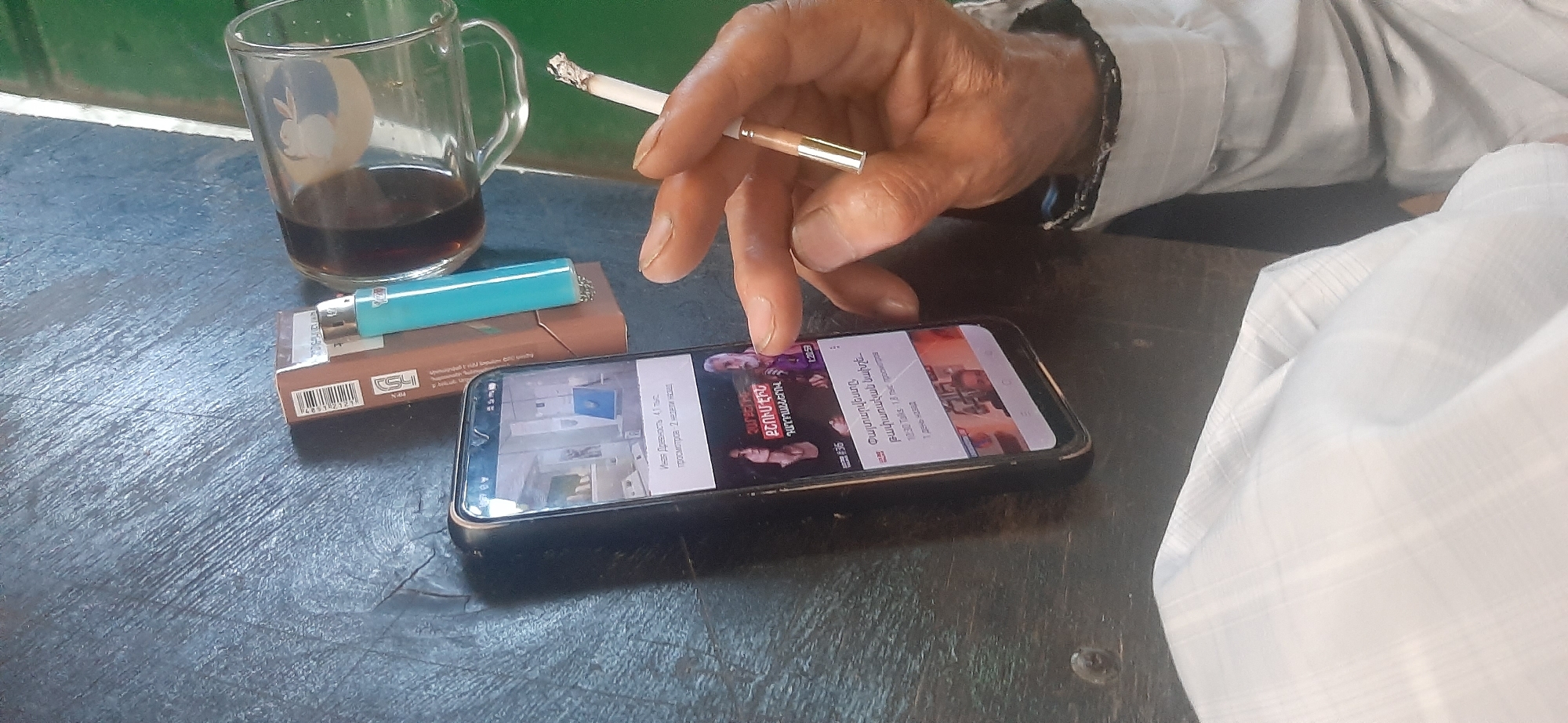 An overhead, close-up shot shows a person's hands and a smartphone on a dark, textured surface, likely a table. The person, visible from the chest down, is wearing a light-colored, long-sleeved shirt. One hand holds a lit cigarette between the index and middle fingers, with smoke rising from the tip. The other hand is reaching towards the smartphone, which displays a YouTube video with Armenian text. Beside the phone is a pack of cigarettes and a light blue lighter. To the left, a clear glass mug filled with a dark liquid, possibly coffee or tea, sits on the table. The background is out of focus, showing a hint of green. The scene appears to be indoors, with natural light illuminating the subjects.