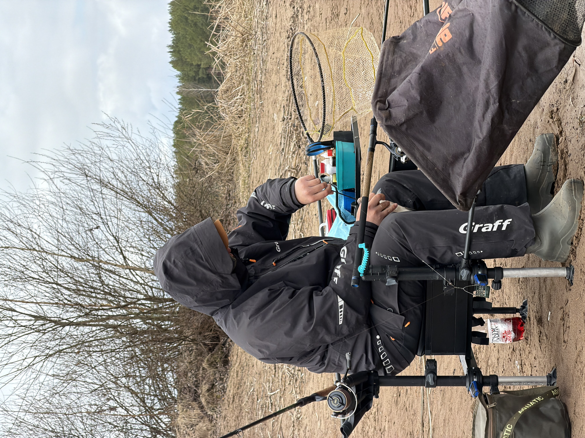 A person dressed in dark, hooded fishing gear, including a jacket and pants branded "Graff" and grey rubber boots, is seated on a black fishing chair or station on a sandy river or lake bank in Chaykovsky, Russia. The individual is actively engaged in what appears to be preparing fishing tackle or bait, with their hands focused on items within a multi-compartment fishing box.
The fishing station is equipped with various tools and containers, including small bottles and what looks like bait. A red bottle with Cyrillic text "ТОРЫХ" (TOROKh) is visible, attached to the side of the station. A fishing rod with a reel is set up, extending into the foreground. To the right of the person, a yellow mesh keepnet is coiled and positioned near other fishing equipment, including a dark bag branded "AQUATIC" resting on the ground.
The setting is outdoors, likely during late winter or early spring, characterized by a sandy or muddy bank with patches of dry grass. Behind the person, a large deciduous tree with bare branches stands against an overcast sky. In the distance, a line of evergreen trees forms the backdrop. The weather appears cool and cloudy. The scene depicts a focused and practical moment of a fishing activity.