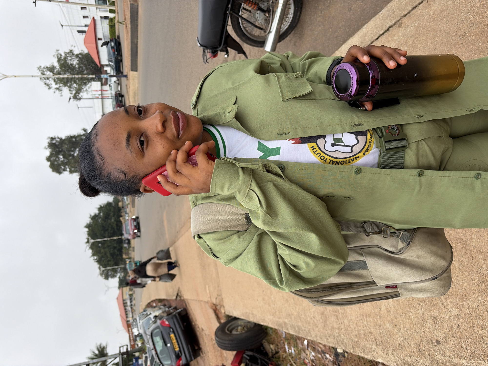 A young woman, identified as a National Youth Service Corps (NYSC) member, stands on a sidewalk in Jos, Nigeria, engaged in a phone conversation. She is wearing the standard NYSC khaki uniform, consisting of a long-sleeved shirt and trousers, with a white t-shirt featuring the NYSC emblem visible underneath. A khaki-colored backpack is slung over her left shoulder, and she holds a metallic water bottle with a purple cap in her right hand.
The background reveals a street scene characteristic of Jos. Cars, including a black Peugeot sedan, are parked along the side of the road. A motorcycle is parked nearby, and pedestrians can be seen walking in the distance. The architecture suggests a mix of commercial and residential buildings, with some sporting red roofs. The sky is overcast, indicating a cloudy day. The sidewalk she stands on appears to be made of concrete, with some signs of wear and tear. Debris is visible on the ground near a damaged vehicle on the left side of the frame. The overall impression is that of an urban environment in Nigeria, with everyday activities unfolding.