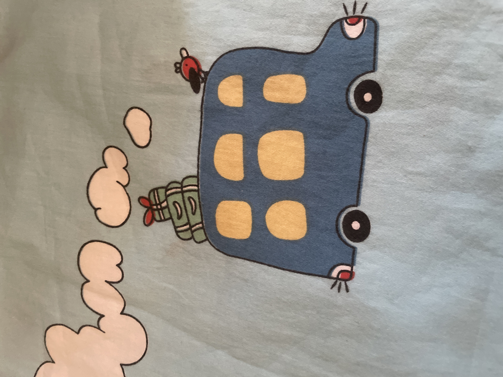 The image shows a cartoon drawing on a light blue fabric. The drawing depicts a blue bus with yellow rectangular windows. On top of the bus, there are stacked green boxes with a red ribbon. A small red bird with black wings is perched on the roof of the bus. To the left of the bus, there are three stylized clouds in off-white with black outlines. The fabric appears to be a textile, possibly part of clothing or home decor. The image is a close-up, and no background elements beyond the drawing and the fabric are visible. There is no indication of the location in Tashkent, Uzbekistan.