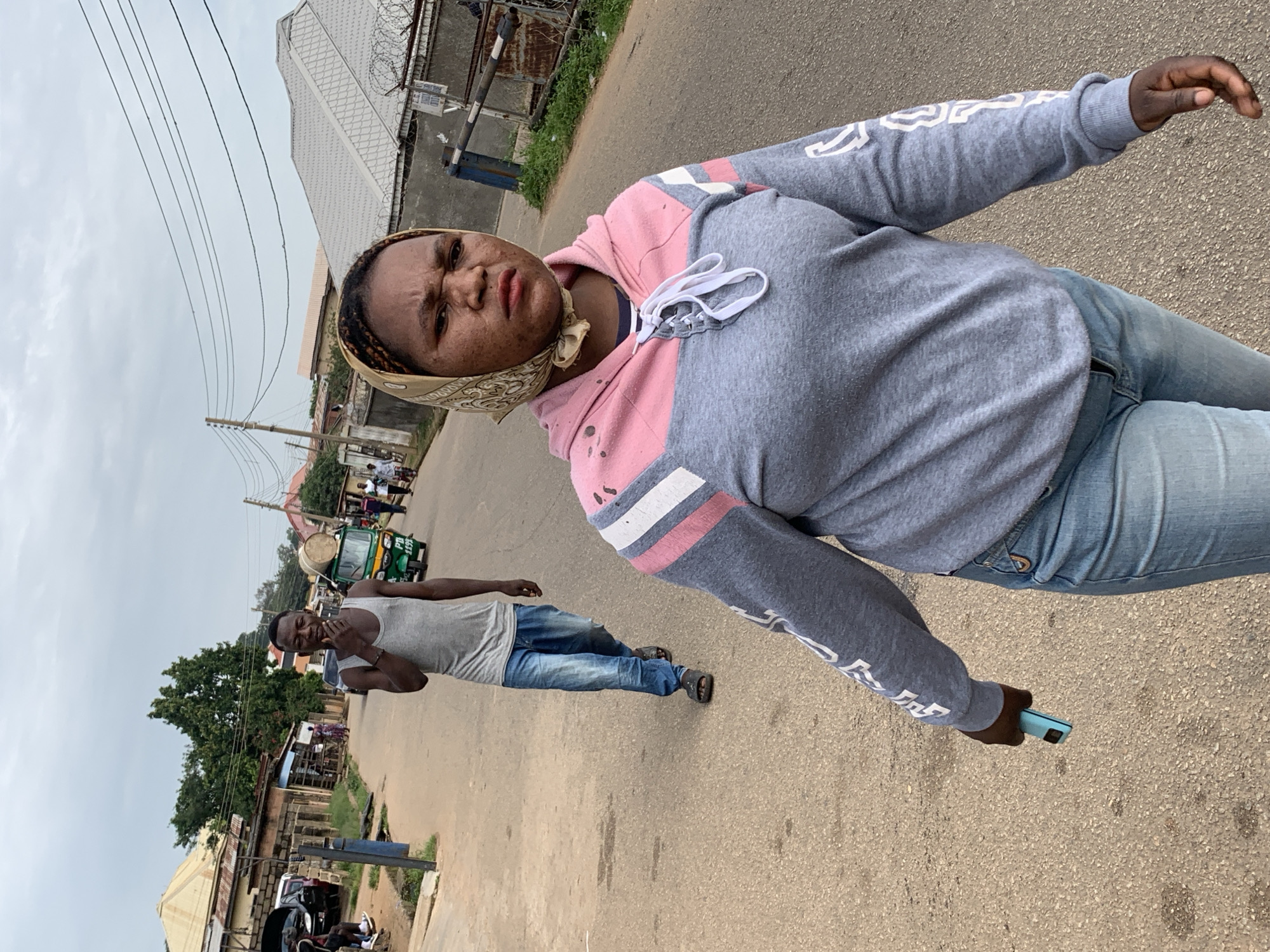 The image depicts a street scene in Jos, Nigeria, likely in a residential or market area. In the foreground, a woman wearing a grey and pink hoodie and light blue jeans walks towards the camera. She has a patterned headscarf tied around her head and is holding a light blue mobile phone in her right hand. Her facial expression is tense, and she has visible skin blemishes. Behind her, a man in a grey tank top and blue jeans walks away from the camera, smoking and carrying a large cylindrical object, possibly a drum or barrel, on his head, balanced on a rack attached to a small motorized tricycle. The tricycle is green and has a sign with "PB 1175" written on it. The street is paved with asphalt and has a slight incline. Buildings line both sides of the street, with some appearing to be shops or houses. Power lines are visible overhead against an overcast sky. The overall environment suggests a bustling, everyday urban setting.