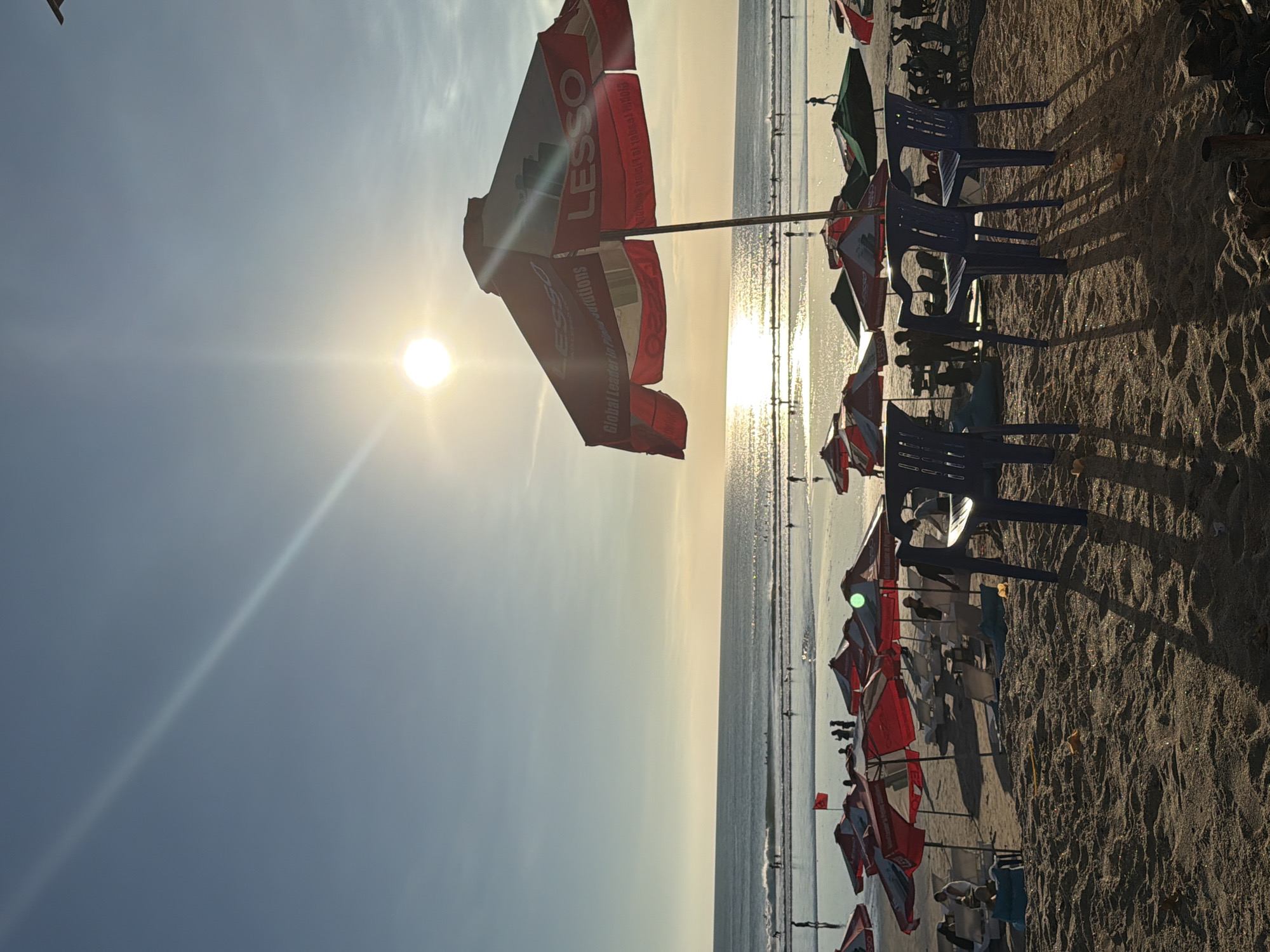 The image captures a serene beach scene in Kuta, Indonesia, during what appears to be late afternoon or early evening. The sun, low in the sky, casts a warm glow, creating a bright reflection on the calm ocean waters. Several beach umbrellas, predominantly red and white, are set up on the sandy shore, offering shade to beachgoers. Some of these umbrellas are branded with the word "LESSO". In the foreground, blue plastic chairs are arranged on the sand, with long shadows stretching out from them. People are visible scattered across the beach and in the shallow water, some wading, others walking, and a few appearing to be surfing in the distance. The overall atmosphere is peaceful and relaxed, typical of a popular beach destination.