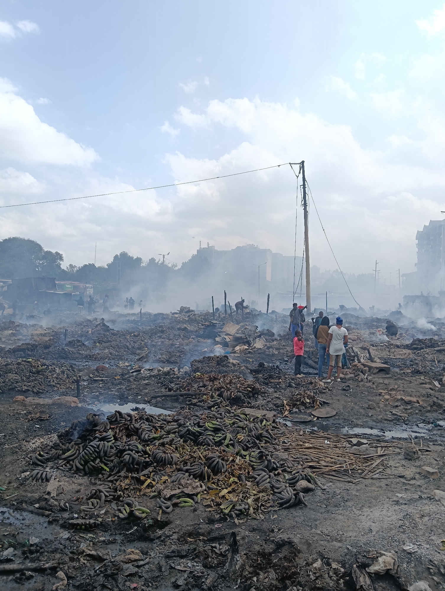 The image depicts a scene of destruction and aftermath in what appears to be a market or informal settlement in Nairobi, Kenya, likely following a fire. The foreground is dominated by heaps of burnt debris, including what seems to be a large quantity of damaged bananas, mixed with ash and charred wood. Smoke still lingers in the air, creating a hazy atmosphere that obscures distant structures.
Several individuals are present, standing amidst the destruction. In the middle ground, a group of people, including adults and children, are observing the scene. One person in a pink shirt and blue jeans stands out, looking towards the left. A person in a white shirt and blue jeans is facing away from the camera. Further back, other figures are visible, some appearing to be sifting through the debris.
In the background, damaged structures, possibly market stalls or dwellings, are visible, some with corrugated metal roofs. Beyond these, more substantial buildings, including multi-story structures, can be seen, suggesting an urban environment. A prominent utility pole with wires stands in the mid-ground, a stark reminder of the city's infrastructure amidst the chaos. The sky is partly cloudy, with a light haze, indicative of the residual smoke. The ground is uneven and appears to be composed of dirt, ash, and scattered debris.