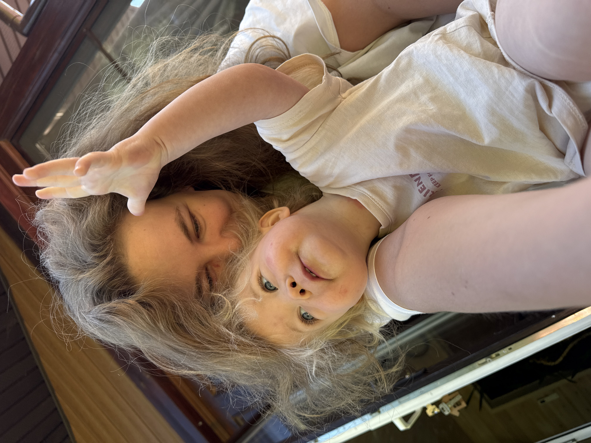 The image captures a close-up portrait of an adult female and a young child in what appears to be an outdoor or porch setting in Копнино, Russia. The adult, with long, wavy, light brown hair, is positioned behind and to the left of the child, with her face partially visible, angled downwards. Her eyes are visible in some crops, and she appears to be looking at the child. Her right arm is around the child. The child, a fair-skinned toddler with blonde, curly hair and blue eyes, is in the foreground, looking upwards and slightly to the right with a soft expression. The child's left hand is raised with fingers splayed, positioned near the adult's head. Both individuals are wearing light-colored, short-sleeved garments; the child's shirt has a faded, reddish-brown text partially visible on the left side, reading "EN" and "KEEP H". The background consists of dark brown wooden structures, likely a house exterior or a door/window frame, with a white door frame and a metallic handle/lock mechanism visible on the far left. The scene is illuminated by natural light.