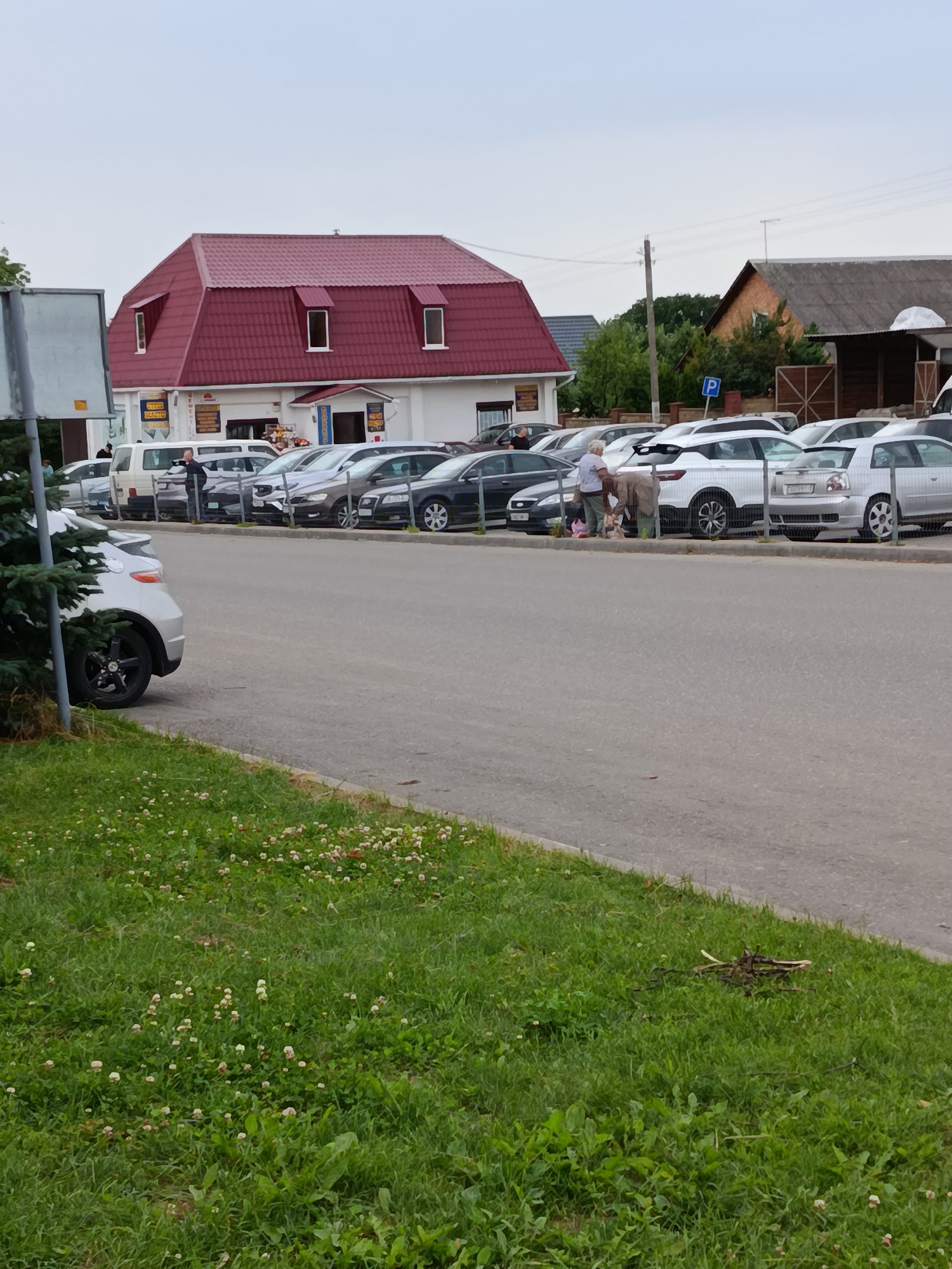 The image depicts an outdoor scene in a parking lot adjacent to a building with a distinctive red mansard roof. Several cars are parked in rows. In the mid-ground, two individuals are interacting near a white car and a light-colored sedan. One person appears to be bending down, possibly retrieving items from a bag, while the other stands nearby. A building with multiple windows and a prominent red roof is visible on the left side of the image, and other buildings and trees are present in the background. A parking sign with a 'P' symbol is visible on a pole. The foreground consists of a paved road and a grassy area with small white flowers. The sky is overcast. The scene is consistent with a commercial or residential area in Смілавіцкі сельскі Савет, Belarus.