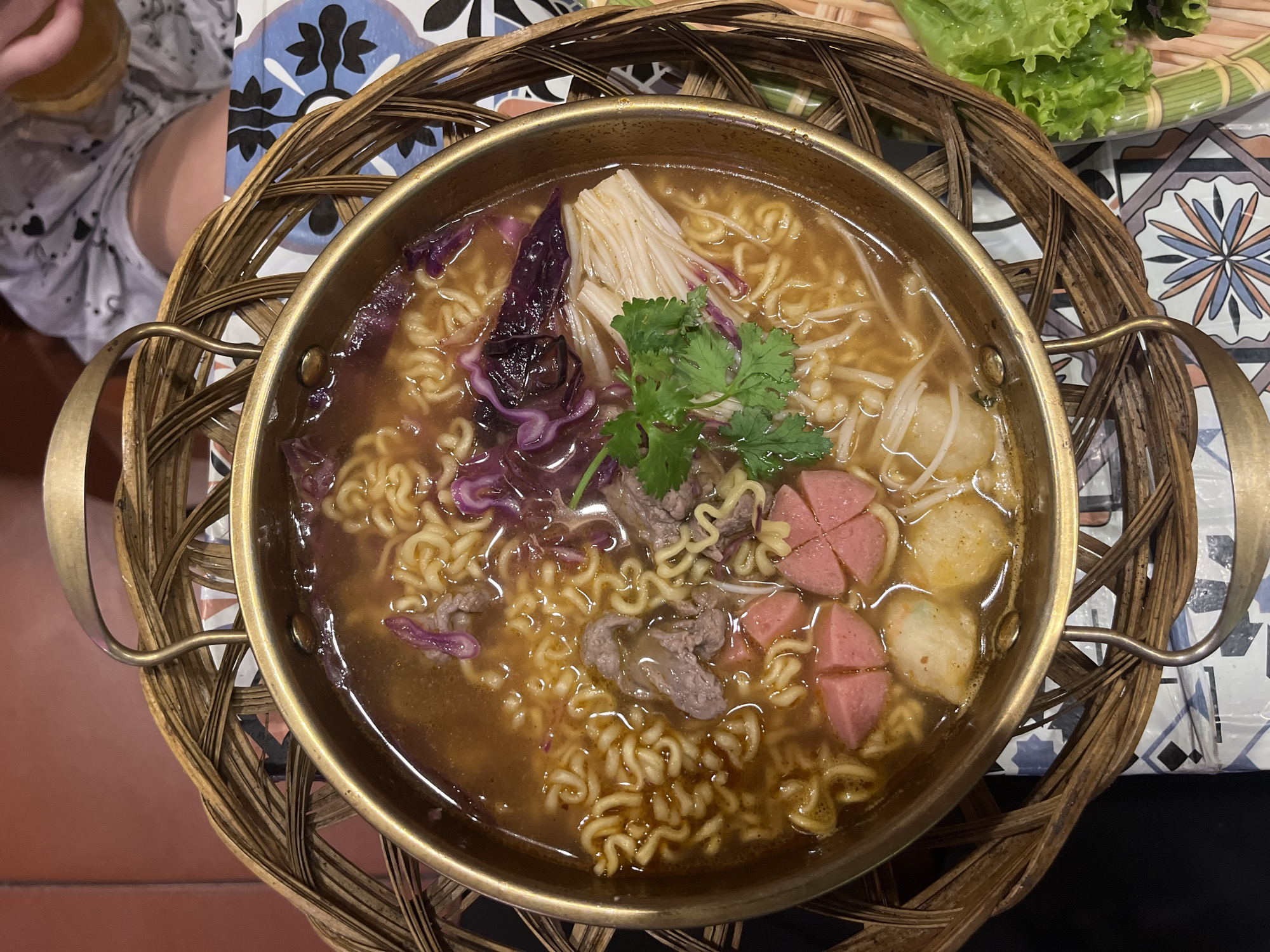 A close-up, overhead shot captures a steaming bowl of noodles, served in a metallic pot with ornate woven handles, nestled within a larger woven basket. The bowl contains a rich broth filled with wavy noodles, thinly sliced beef, pink-hued sausage segments, white fish balls, and delicate strands of enoki mushrooms, all garnished with fresh green cilantro and vibrant purple cabbage. To the right, a bundle of crisp green lettuce sits beside the bowl. The background reveals a patterned tiled surface and a hint of a person's torso, suggesting a dining setting. This scene is located in the city of Bac Nha Trang Ward, Vietnam.