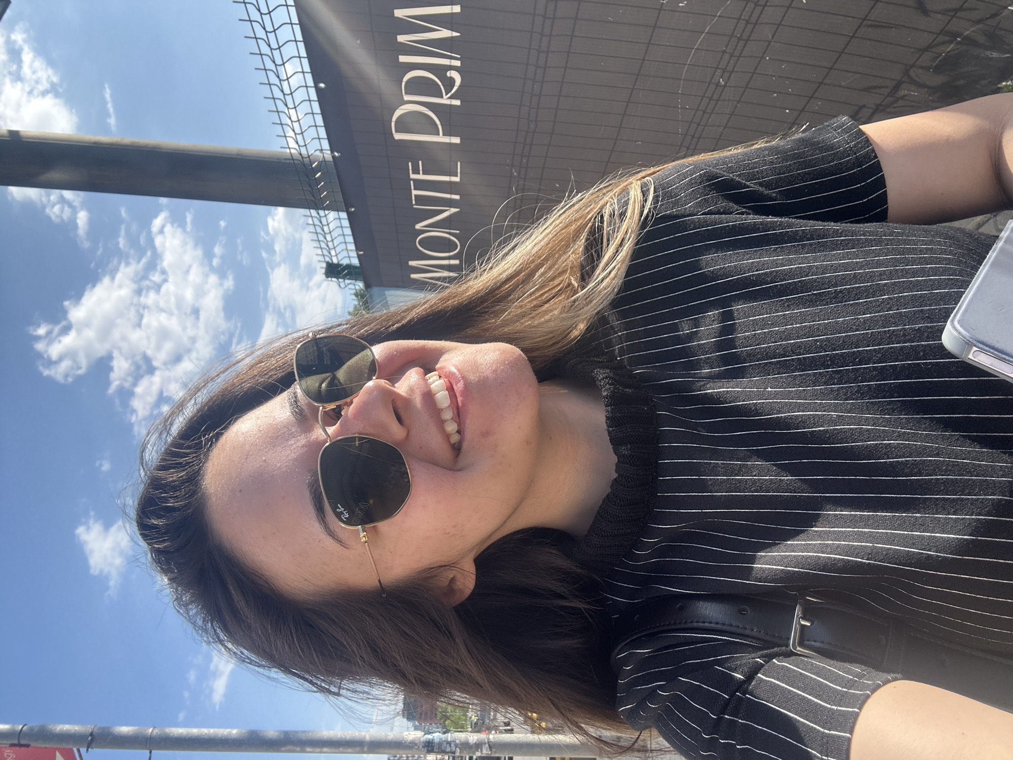 A woman in sunglasses stands in front of a sign that reads "MONTE PRIMA" in Sofia, Bulgaria. The woman is wearing a black and white striped shirt. The sky is blue with scattered clouds. A black strap is visible. A grey metal structure is also visible.