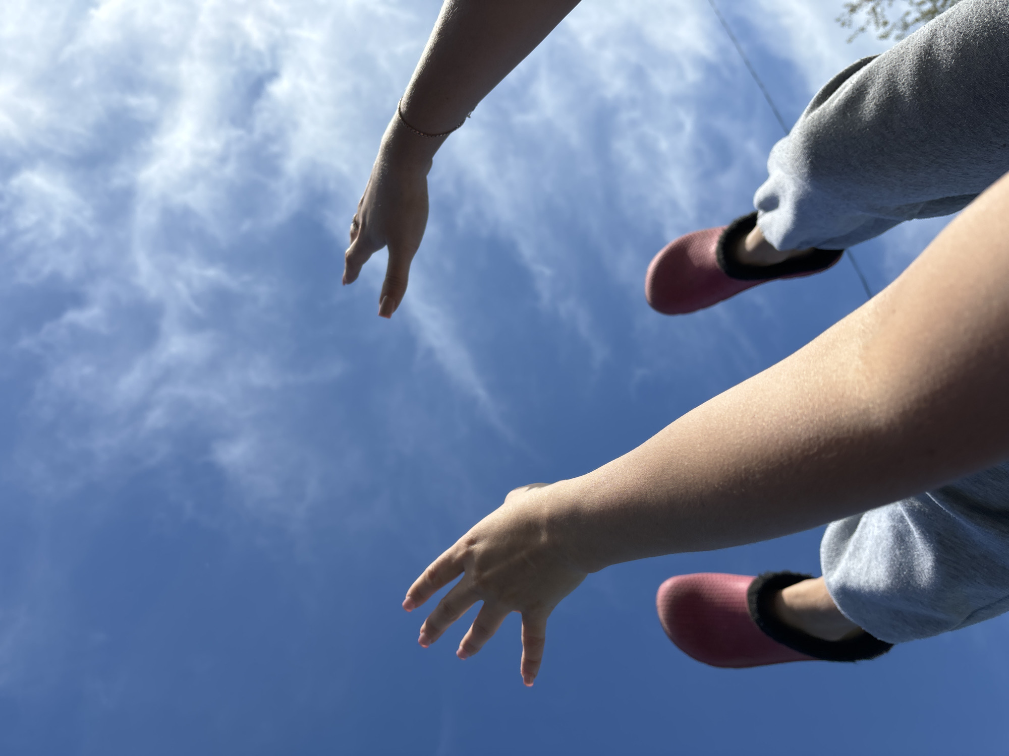 The image is a low-angle shot taken outdoors in daylight, looking up towards the sky. The sky is blue with scattered white clouds. Two arms and legs are visible in the frame, suggesting the perspective of someone lying on the ground looking up. One arm, adorned with a thin bracelet, extends from the upper left corner. The other arm is positioned in the lower right corner. The legs, clad in gray sweatpants and pink slippers with dark trim, are also visible near the right side of the frame. There is a faint outline of a tree at the top right of the image. It is not possible to verify the location as Chaikovsky, Russia from this image.