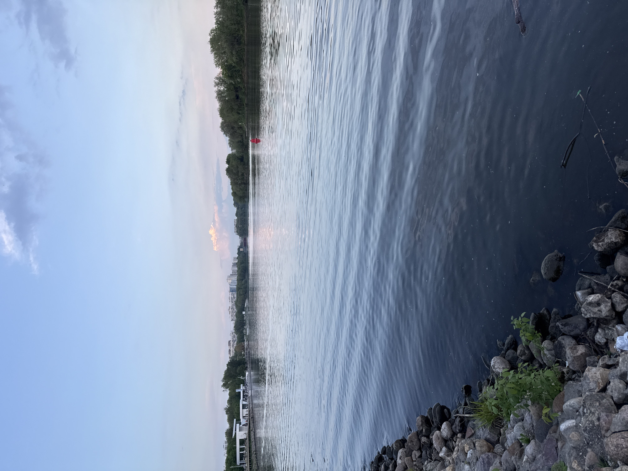 This image depicts a view of a body of water, likely a river or lake, in Moscow, Russia. The scene is captured at dusk or dawn, judging by the soft light and the colors in the sky. The water is rippled, reflecting the sky and the distant cityscape. Along the shoreline, there are rocks and some vegetation. In the distance, buildings and trees can be seen along the horizon. A red buoy or marker is visible on the water. The sky is partly cloudy with shades of blue, grey, and faint orange. There are no people visible in the image.