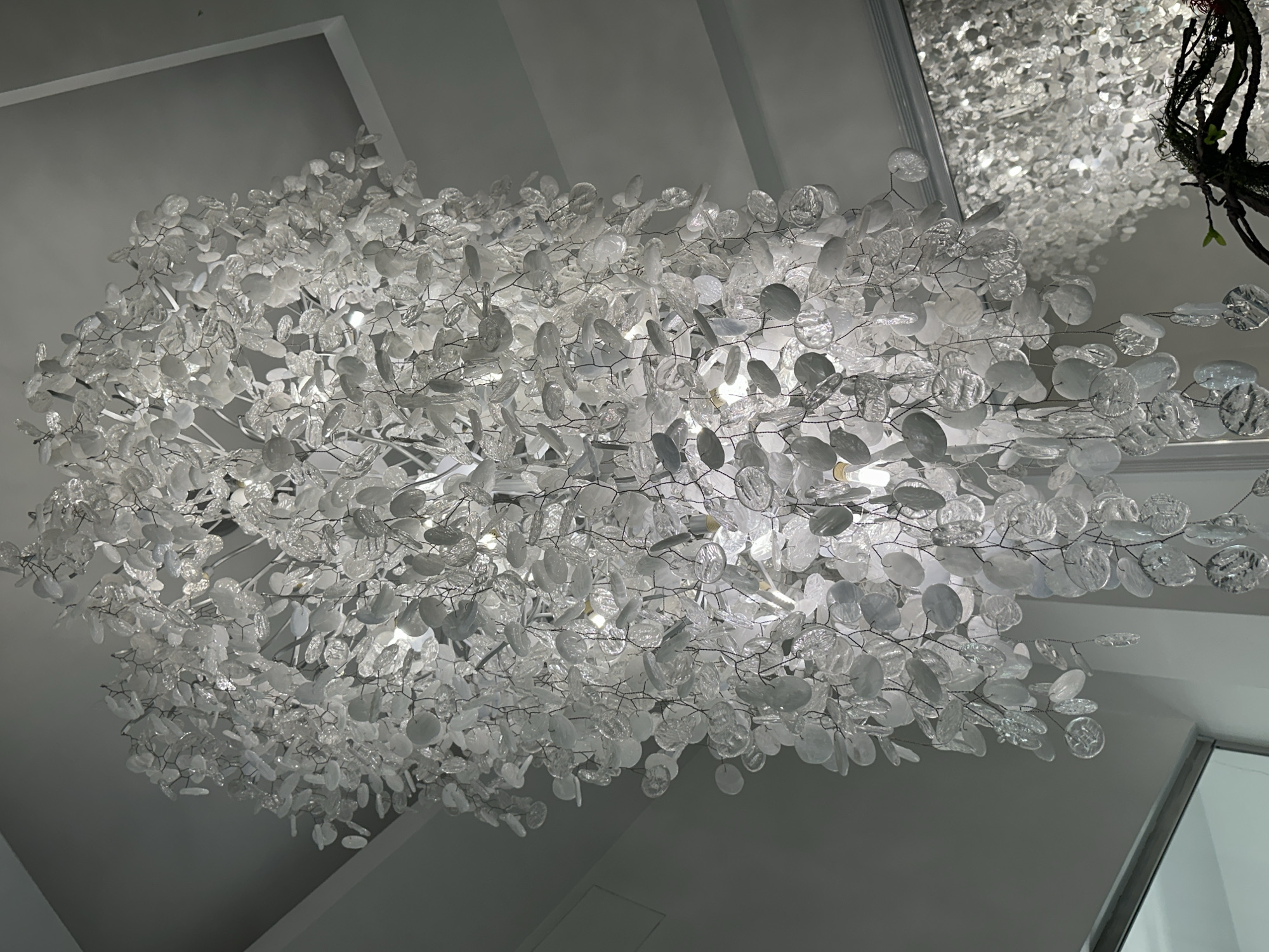 The image shows a large, decorative chandelier hanging from a white ceiling. The chandelier appears to be composed of numerous small, white, circular or oval-shaped translucent elements attached to a metal framework. The lighting fixture is positioned prominently in the center of the frame. Another chandelier is visible in the upper-right corner, reflected in a mirror, or a surface that appears reflective. Parts of the surrounding architecture (ceiling, walls) are white or light gray. The image does not contain sufficient information to determine the location in Khimki, Russia.