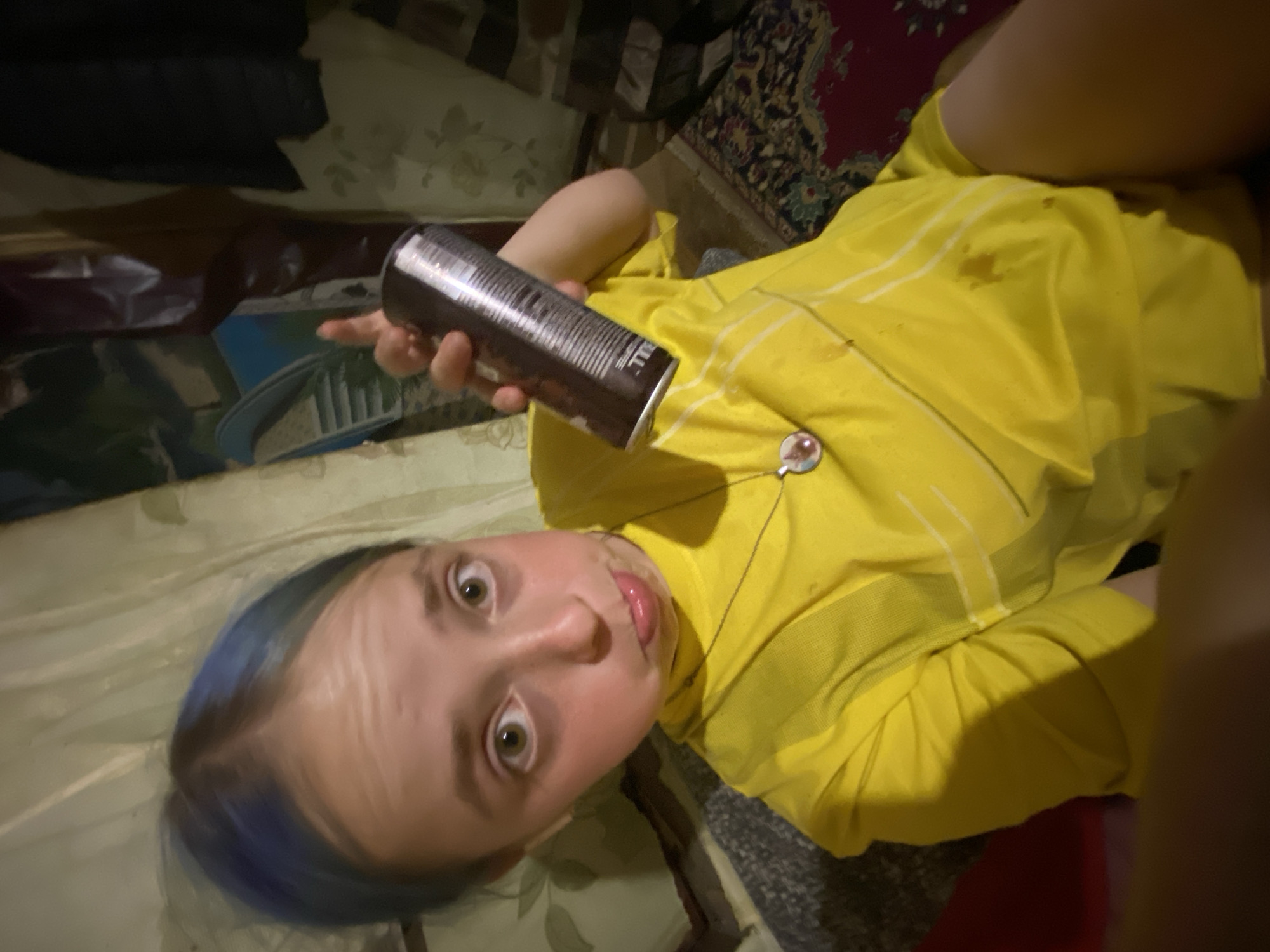 In this media file, a young person with blue-tinted hair is lying down, holding a dark-colored can in their right hand. They are wearing a bright yellow t-shirt with thin white stripes. A small, circular pendant hangs from a necklace around their neck, resting on the t-shirt. The person's face is close to the camera, and they appear to be looking directly at it with wide eyes and their tongue slightly sticking out, suggesting a playful or surprised expression. The background is dimly lit and appears to be an indoor setting with what looks like a patterned rug and possibly furniture. The overall impression is casual and informal, possibly taken in a home environment in Nova Praha, Ukraine. No specific time of day or weather conditions are discernible. The text on the can is not clearly legible due to the resolution and angle.