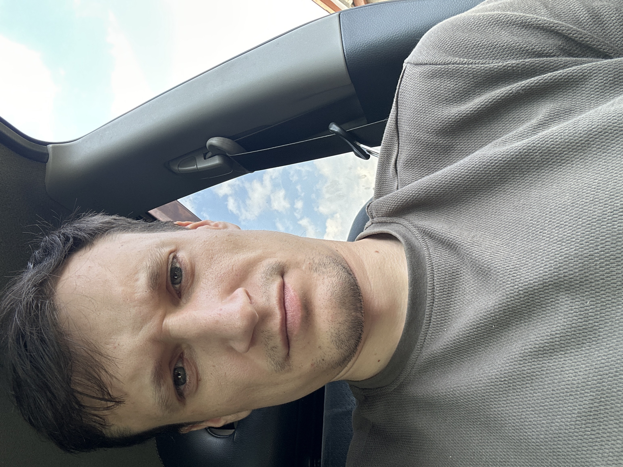 The image shows a close-up selfie of a man inside a vehicle. The man has dark hair and is wearing a grey t-shirt. He is looking directly at the camera with a neutral expression. The interior of the car is visible, including the headrest and part of the car's pillar. Through the car window, a cloudy blue sky is visible. The location is the city of Makiv, Ukraine.