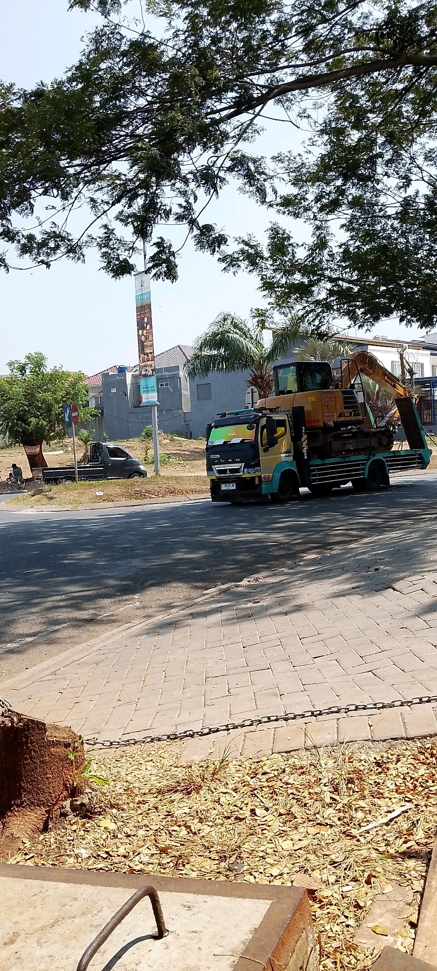 The image depicts a street scene in Gunung Putri, Indonesia, under a clear sky and dappled sunlight filtering through a large tree canopy. A yellow and green flatbed truck carrying an excavator is driving on an asphalt road. The truck has a black and chrome front grille with the "FURO" logo. The excavator is orange and yellow with "SANY" and "JX140" visible on its arm. On the left side of the road, a dark-colored pickup truck is parked, and a person is sitting on a bicycle nearby. Further back, houses with gray walls and tiled roofs are visible, along with palm trees and other vegetation. A tall banner with the text "ECOVIA" and "CYDORA" is also present. The foreground shows a paved sidewalk with a metal chain and a concrete structure with a rusty metal handle. The ground is covered with dry leaves and scattered debris. The overall impression is of a typical street scene in a residential or developing area.