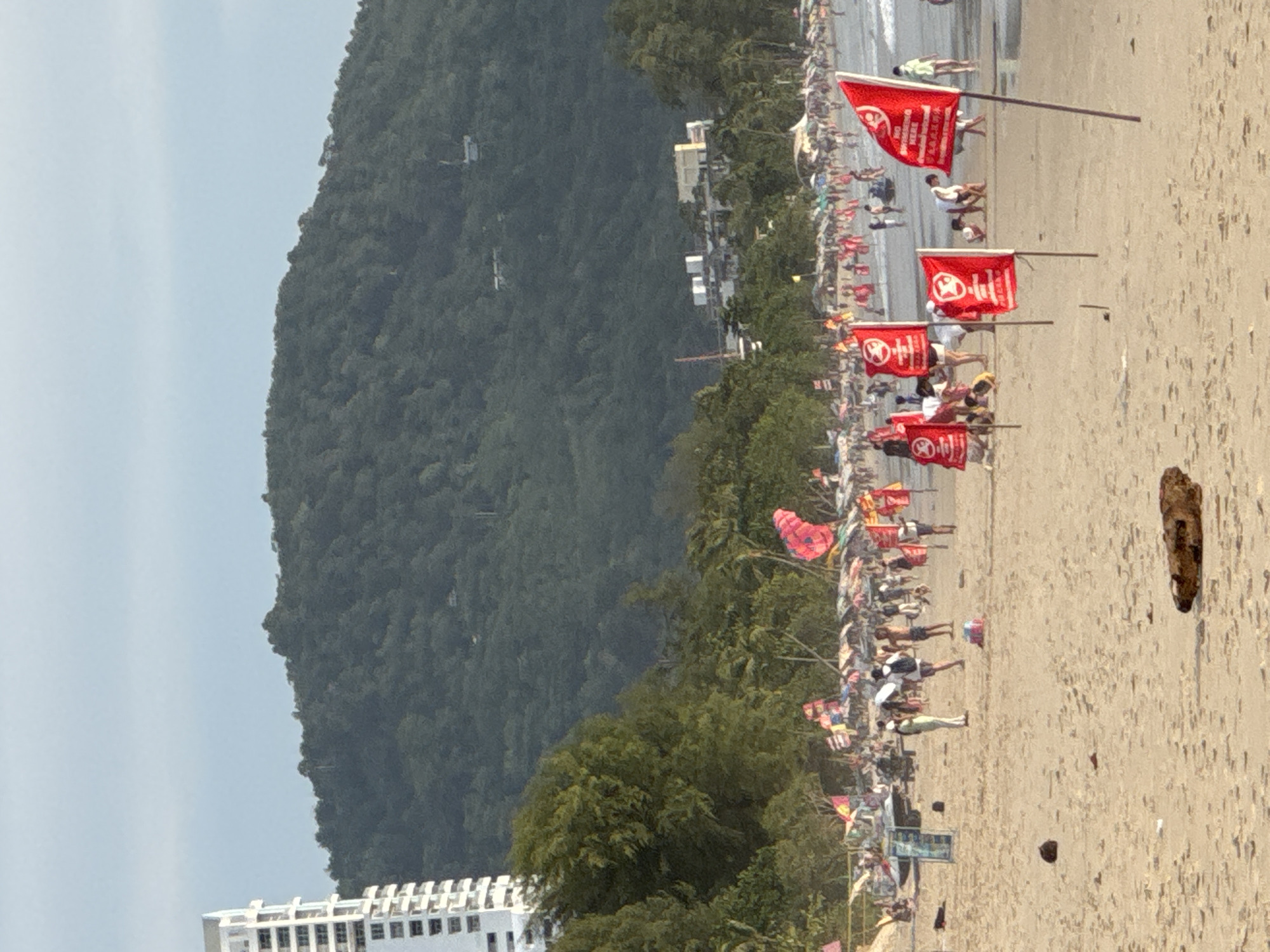 The image depicts a crowded beach scene in Patong, Thailand, with a mountainous, tree-covered backdrop. In the foreground, several red flags are planted in the sand, some with Chinese characters and English text that reads "NO SWIMMING HERE". People are scattered across the beach, some walking, some sitting, and others appearing to be playing or gathering near beach umbrellas. A tall, white building is visible on the left side of the frame, characteristic of the high-rise hotels found in Patong. Further down the beach, a line of umbrellas and people suggests a vibrant atmosphere. The ocean is visible in the background with gentle waves. The overall impression is a busy tourist beach, possibly with advisories against swimming due to ocean conditions.