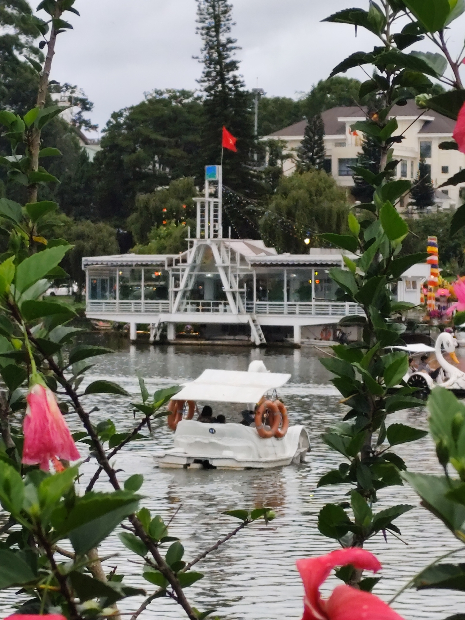 The image depicts a lake in Da Lat, Vietnam, with a white, two-story building on stilts in the background. A red flag is flying atop the building. In the foreground, there are pink hibiscus flowers and green leaves framing the view. A white paddle boat with orange life preservers is floating on the water. In the background, there is a house on the hill and swan-shaped boats. The sky appears overcast.