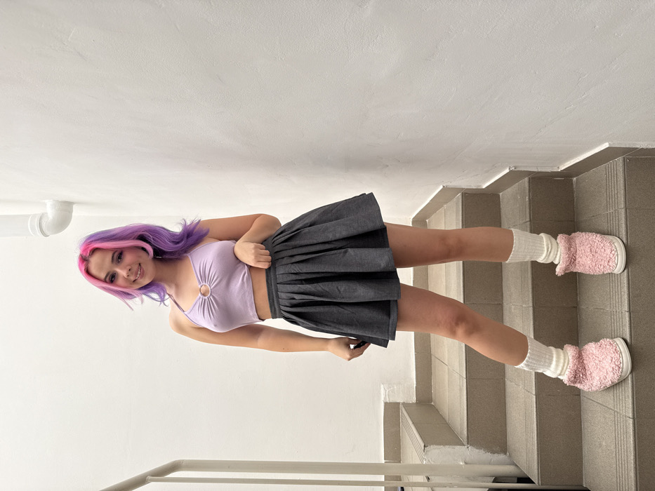 A young adult female stands centrally on a light brown concrete stairwell with white walls. She has wavy hair dyed pink on top and purple underneath, and smiles towards the viewer. She is wearing a light purple halter-neck crop top with a silver O-ring at the chest, a dark grey pleated mini skirt, white ribbed leg warmers, and pink fluffy slipper-style shoes with white soles. Her left hand is near her left hip, touching the skirt, with a dark object partially visible in her grasp. A white metal handrail is positioned on the left side of the stairs, and a white pipe runs vertically along the upper right white wall. The specific location within the City of Zagreb, Croatia, is not discernible from the image details.