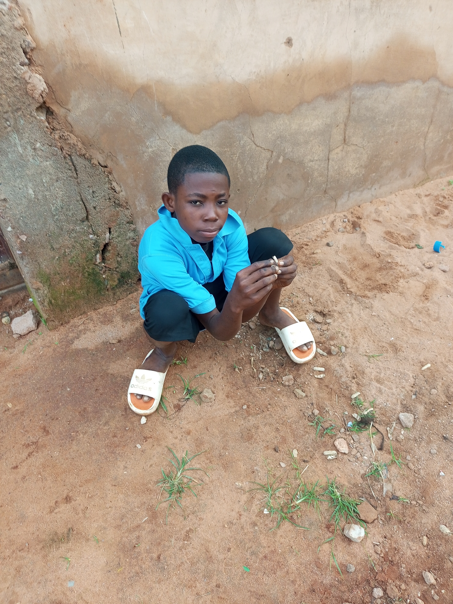 A young boy, appearing to be of African descent and around 8-10 years old, is crouched on sandy ground in front of a weathered, light-colored wall. He is wearing a bright blue collared shirt and black shorts, with white Adidas slides on his feet. His gaze is directed at the viewer. His hands are occupied with a small object, which he holds between his fingers. The ground is a mix of reddish-brown sand with scattered stones and sparse green vegetation. To the left of the boy, a rough, concrete structure is visible, partially obscured by shadow. A small, blue plastic object is visible on the sand in the background, to the right. The overall setting appears to be an outdoor, informal area.
