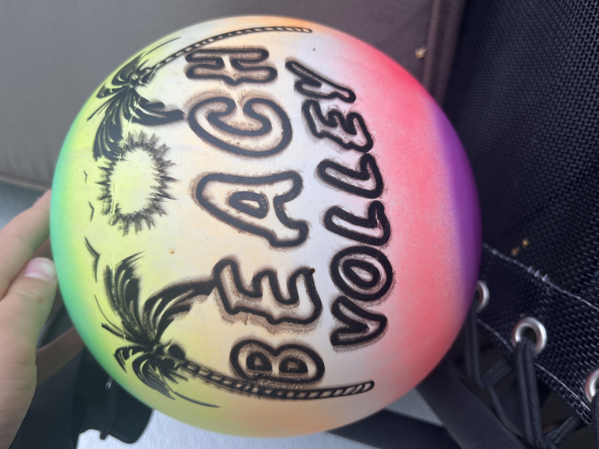 A close-up, eye-level shot shows a beach volleyball with a gradient of green, yellow, pink, and purple. The ball is decorated with black silhouettes of palm trees, a sun with rays, and two birds in flight. Large, black, outlined letters on the ball read "BEACH VOLLEY." A person's hand is visible at the top of the frame, reaching towards the ball. The ball rests on a black mesh surface, possibly the seat of an outdoor chair, with metal grommets and black laces visible in the foreground. The background is blurred, suggesting an outdoor setting. The location is identified as Haabneeme alevik, Estonia.
