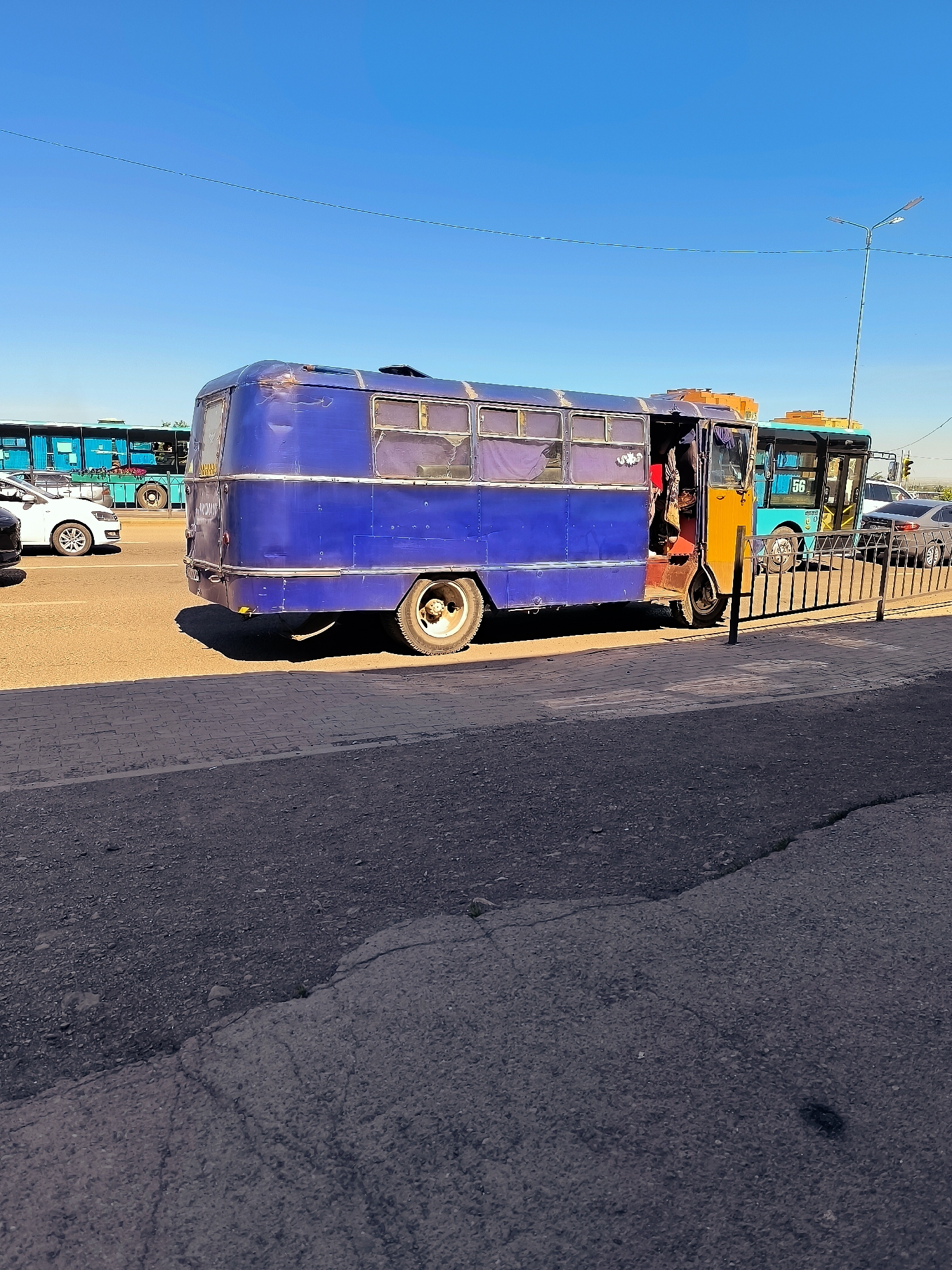 A public bus, painted bright blue with yellow accents, stands on a city street in Qarağandı, Kazakhstan. The bus is old and appears to be in disrepair, with several windows broken or missing. A person is visible inside the open doorway of the bus. In the background, another bus with the number "56" on its side and other vehicles are parked. The street is paved and there is a sidewalk with a metal fence. The sky above is clear and blue, indicating a sunny day. The overall scene depicts a typical street in an urban environment.