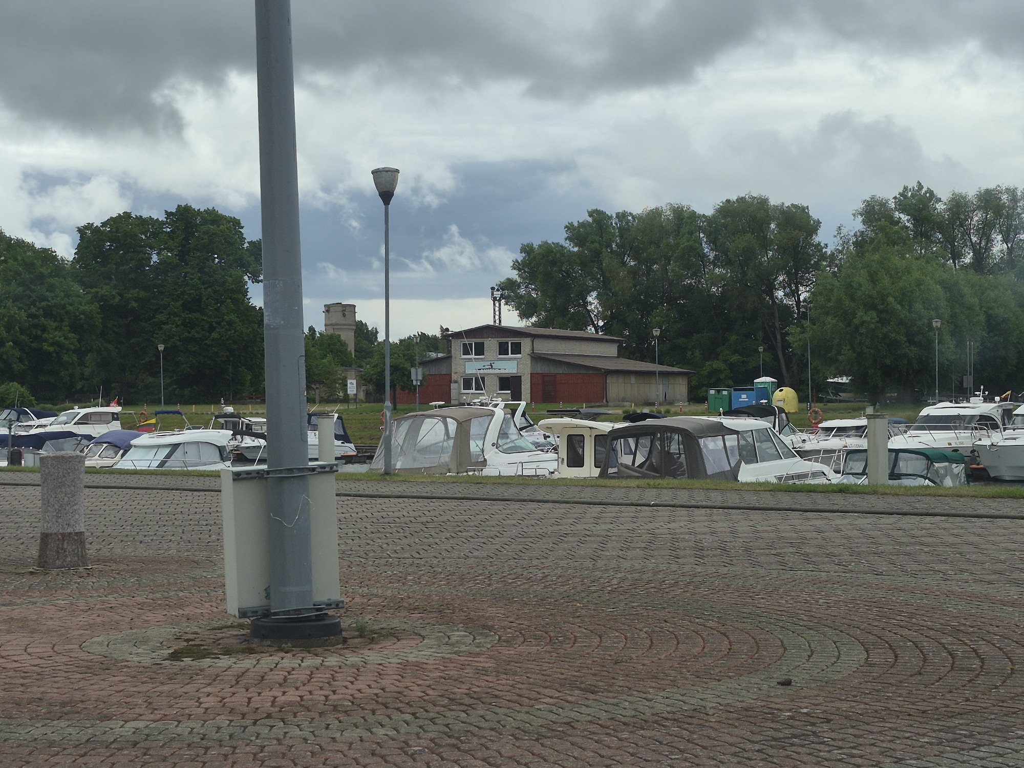 The image depicts a harbor in Šilutė, Lithuania, with several boats docked. The foreground shows a cobblestone walkway with a tall gray utility pole. In the background, a cluster of buildings, including what appears to be a boathouse or marina facility, is visible. Trees line the background, and the sky is overcast with dramatic clouds. The boats are a mix of recreational vessels, with several featuring white hulls and canvas coverings. The overall atmosphere is that of a typical harbor scene on a cloudy day.