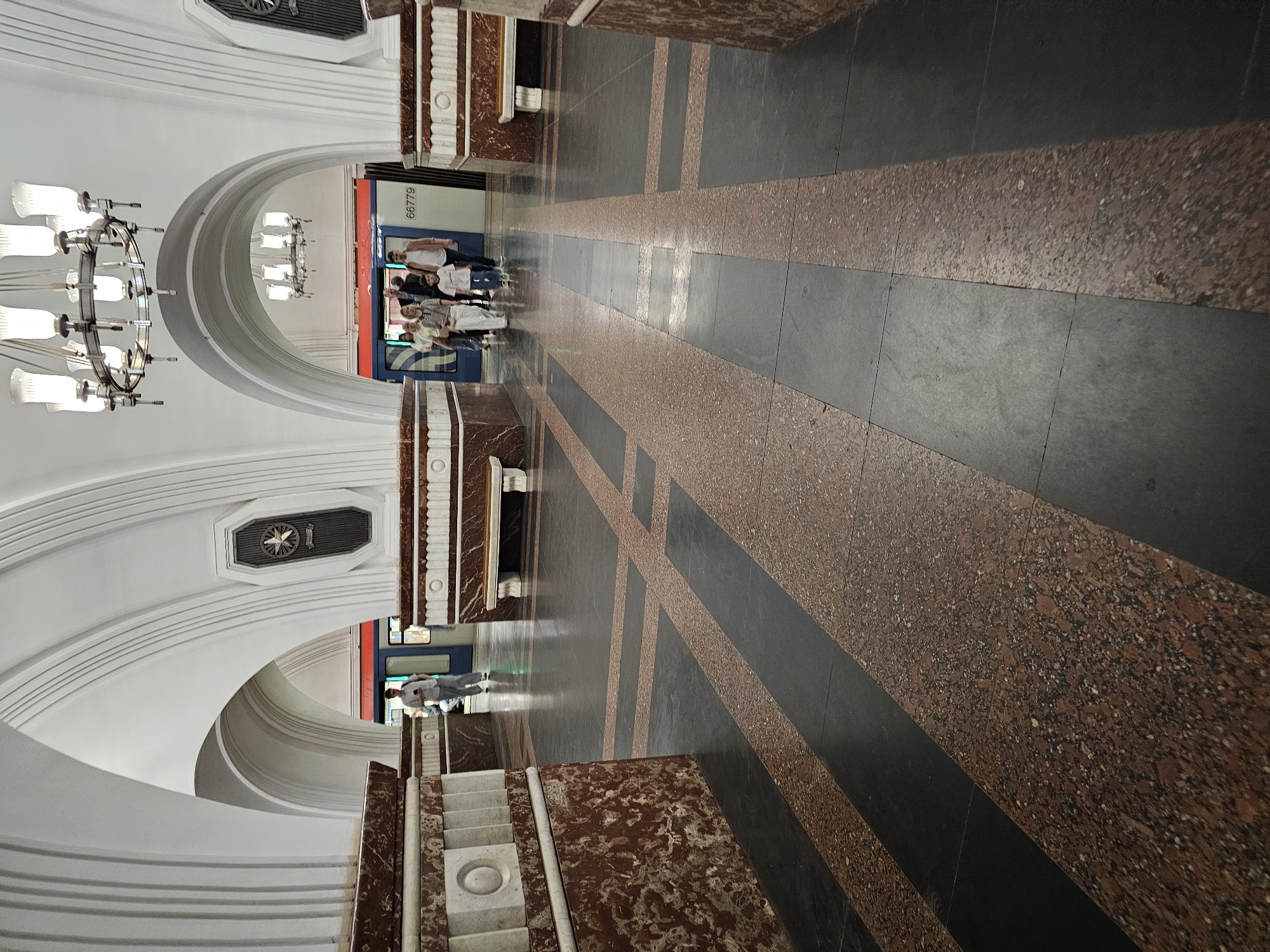 The image shows the interior of the Kiyevskaya metro station in Moscow, Russia. The camera faces down the corridor, showing the floor tiled with alternating bands of dark and red marble. The walls are decorated with white arches and ornate marble panels. Several people stand in the background near a train car. A chandelier hangs from the arched ceiling.