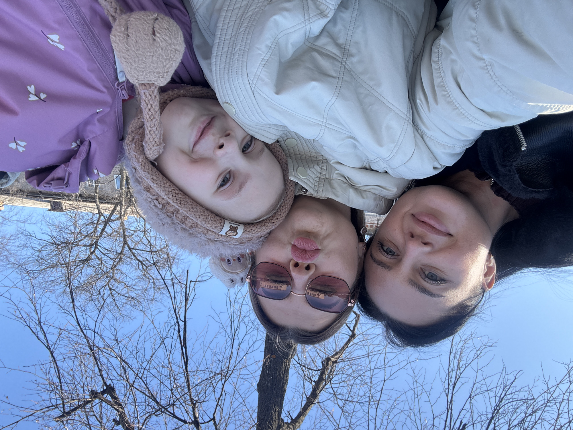 Three individuals are pictured in a selfie taken outdoors, possibly in Chaikovsky, Russia. A young girl with fair skin and blonde hair is on the left wearing a pink hat with rabbit ears and a lavender coat. In the center is a woman with fair skin wearing sunglasses and pursing her lips. On the right is a dark-haired woman smiling slightly. Bare tree branches and a clear blue sky are visible in the background.