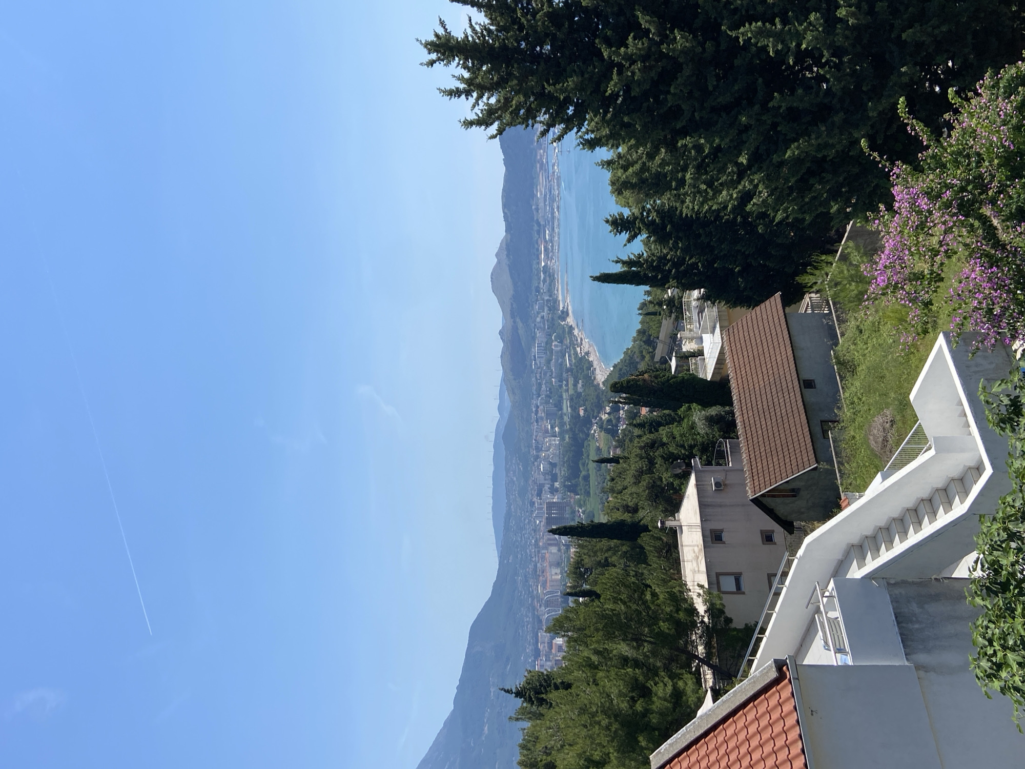 The image depicts a panoramic view of Šušanj, Montenegro, capturing a coastal cityscape nestled between mountains and the Adriatic Sea. The foreground features residential buildings with terracotta tile roofs and white exteriors. A prominent white staircase ascends along the side of one of the buildings. Lush greenery, including tall trees and flowering shrubs with purple blossoms, frames the view. In the midground, the city of Šušanj stretches along the coastline, with buildings and infrastructure visible. The Adriatic Sea extends into the background, bordered by distant mountains under a clear blue sky. A contrail from a passing airplane is faintly visible in the upper left portion of the sky.
