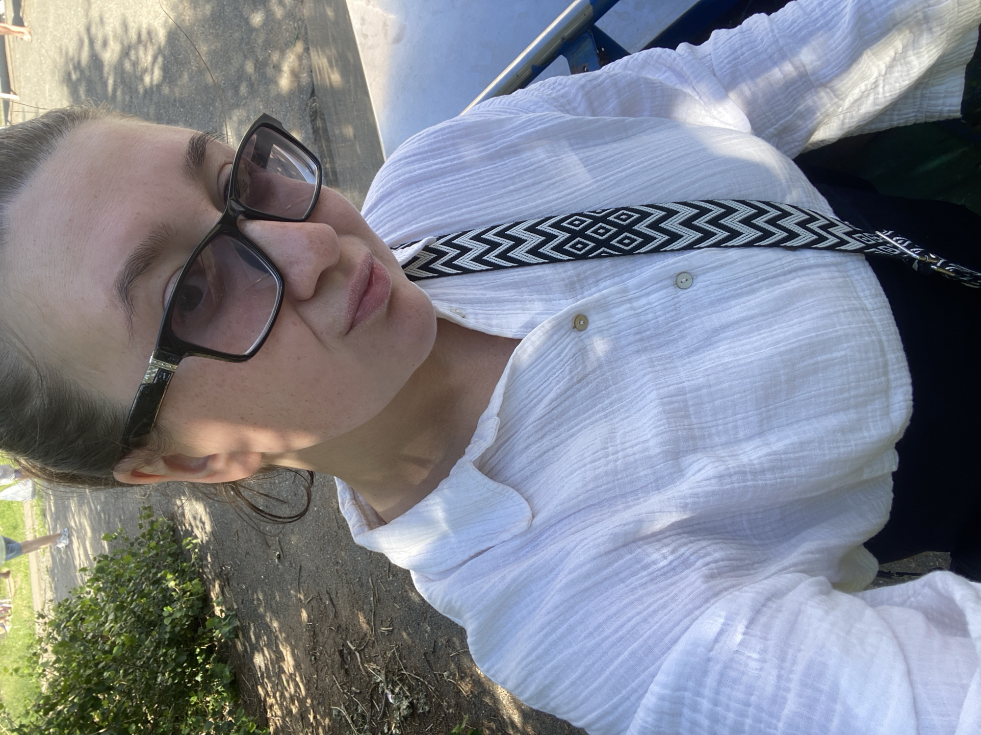 A person is captured in a selfie, their face angled towards the camera. They are wearing black-framed glasses and a white, textured shirt. A patterned strap, in black and white zig-zag and diamond shapes, crosses the shirt from left to right. The person's lips are slightly parted, and their gaze is directed somewhat away from the lens. The background shows a sunlit outdoor scene with dappled shadows from trees on a paved surface. There is greenery in the lower left portion of the image, and a glimpse of another person walking in the distance. The lighting suggests it is daytime, possibly late afternoon given the angle of the shadows. The setting appears to be a park or outdoor recreational area.
