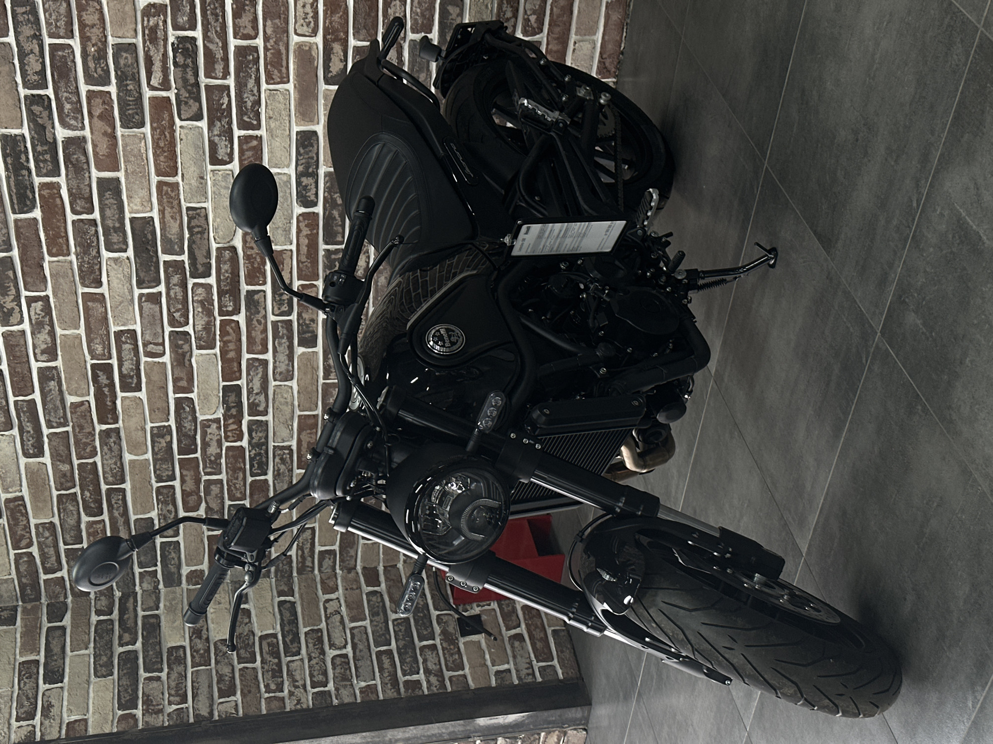 A black Benelli Leoncino 500 motorcycle is stationary, viewed from the front-left, in an indoor setting. The motorcycle rests on its deployed kickstand on a dark grey tiled floor composed of large square tiles. Its features include a round LED headlight, black handlebars, two black rearview mirrors, a black ribbed seat, and knobby-tread tires. A white informational tag, identifying the model as 'Benelli Leoncino 500' and displaying a price (198,900 rubles, partially visible), hangs from the left side of the frame near the engine. The background consists of an interior brick wall with varying shades of brown, red, and beige bricks, delineated by white mortar. A dark vertical structural element is visible on the far left, and a red object is partially obscured behind the front forks. No individuals are present. The location is Rostov-on-Don, Russia.