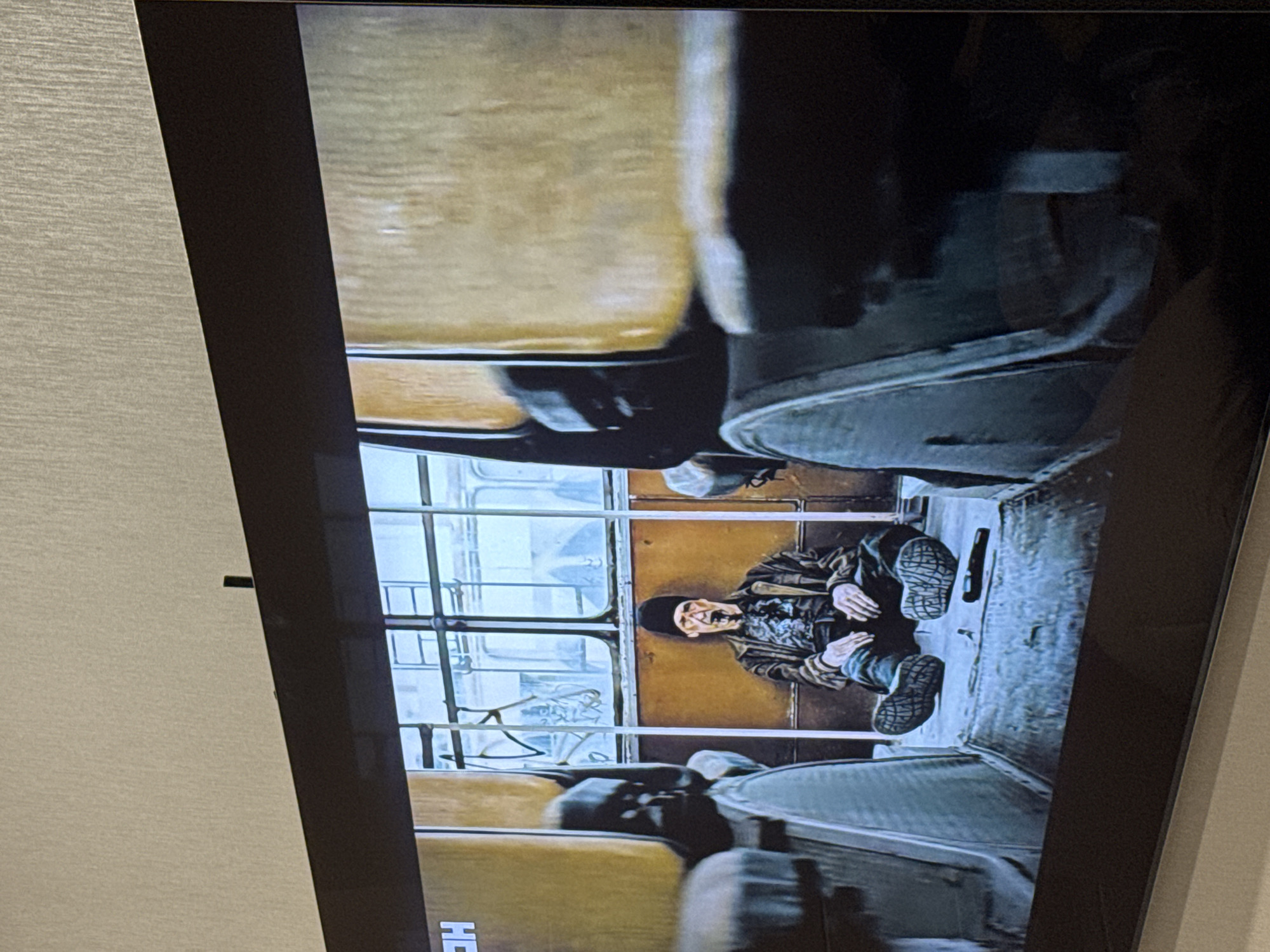 A man is sitting on the floor of what appears to be a tram or bus. He has a black beanie, a patterned shirt, and a jacket. He is sitting between seats with his legs outstretched. The vehicle has a series of windows with a view to the outside, where buildings can be seen. The scene is viewed on a TV screen.