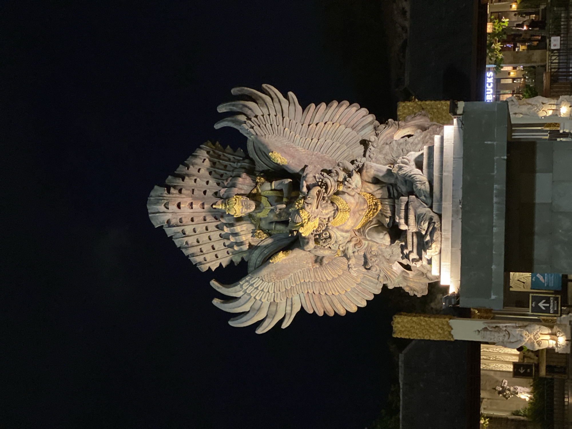 The image captures the Garuda Wisnu Kencana Cultural Park in Kuta Selatan, Bali, Indonesia, at night. The central subject is a massive, ornate statue depicting the Hindu deity Vishnu riding on his mount, Garuda. The statue is illuminated, highlighting its intricate details, including Garuda's feathered wings spread wide and Vishnu seated atop. In the background, a dark sky contrasts with the illuminated statue and the architecture of the park, which appears to include buildings with traditional Balinese elements and signage for a Starbucks. The overall scene suggests a cultural landmark or attraction.