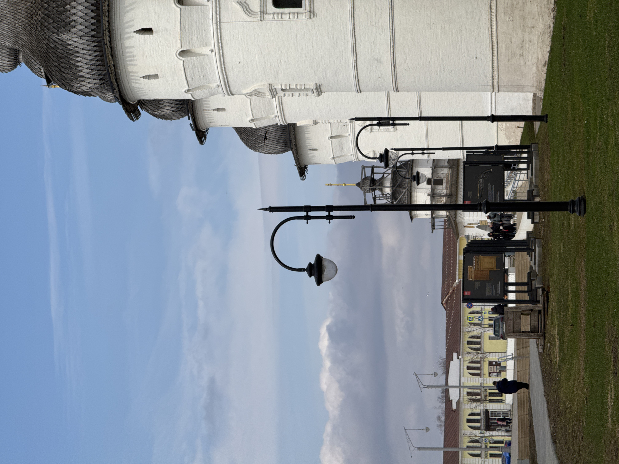 The image depicts a street in Ново-Никольское, Russia. A white brick building with a detailed roof is visible on the upper left. A row of black streetlights runs along the sidewalk. Informational displays stand near the streetlights. People walk along the sidewalk. A yellow building with dark accents is on the opposite side of the street. Patches of green grass border the sidewalk and street. The sky is blue with scattered clouds.