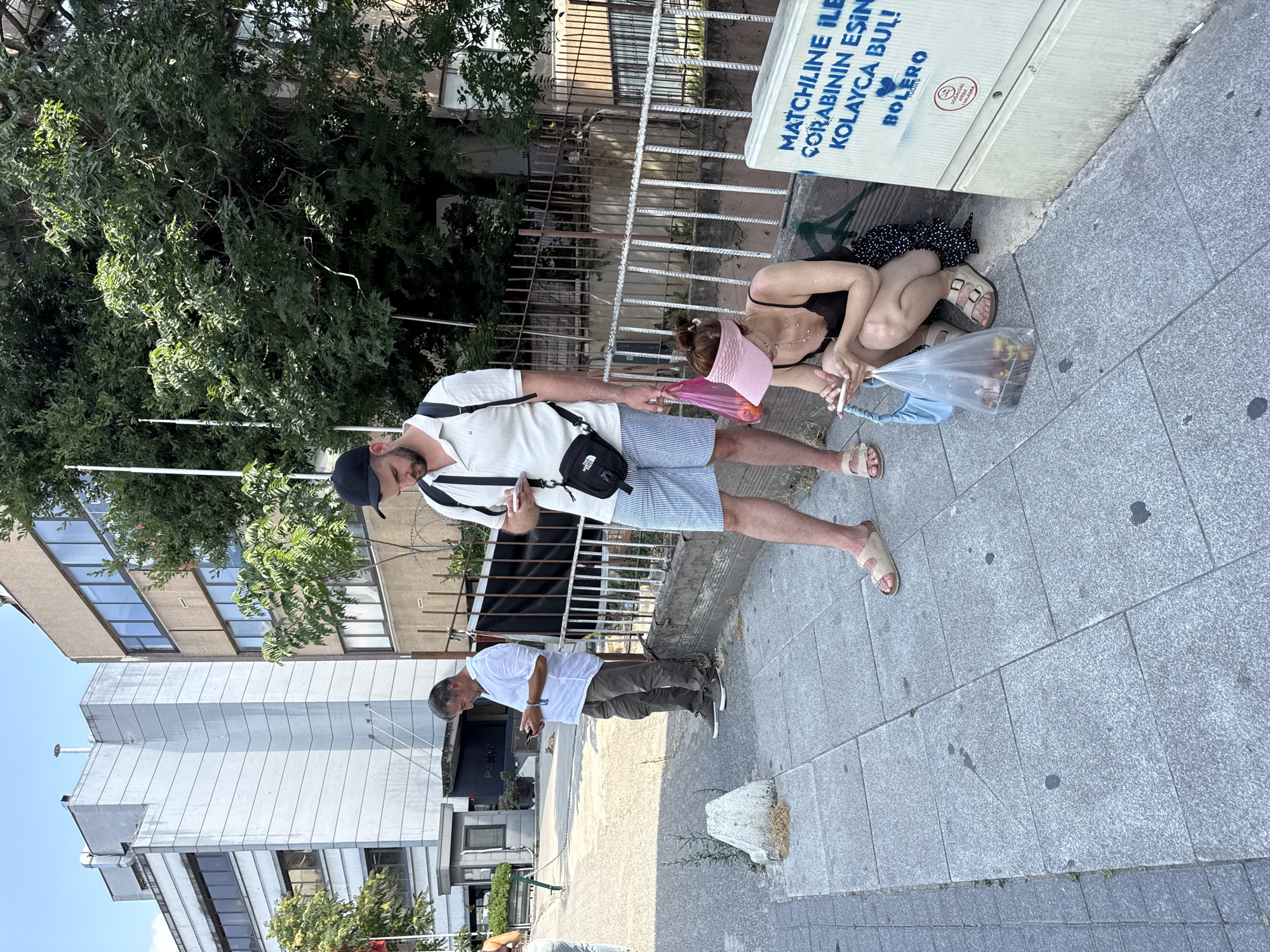 In Kadıköy, Turkey, on a bright, sunny day, three individuals are captured on a paved sidewalk adjacent to a building. A man in a white polo shirt and striped shorts stands holding a mobile phone, a black crossbody bag slung over his shoulder. Beside him, a woman in a black tank top and a pink visor is crouched down, also looking at her phone, with a plastic bag containing food items at her side and a light blue purse on the ground. Further down the sidewalk, another man in a white shirt and gray pants is standing and looking at his phone. The environment includes a metal fence, trees, and a building in the background, with an electrical box on the right side of the frame displaying text in blue spray paint.