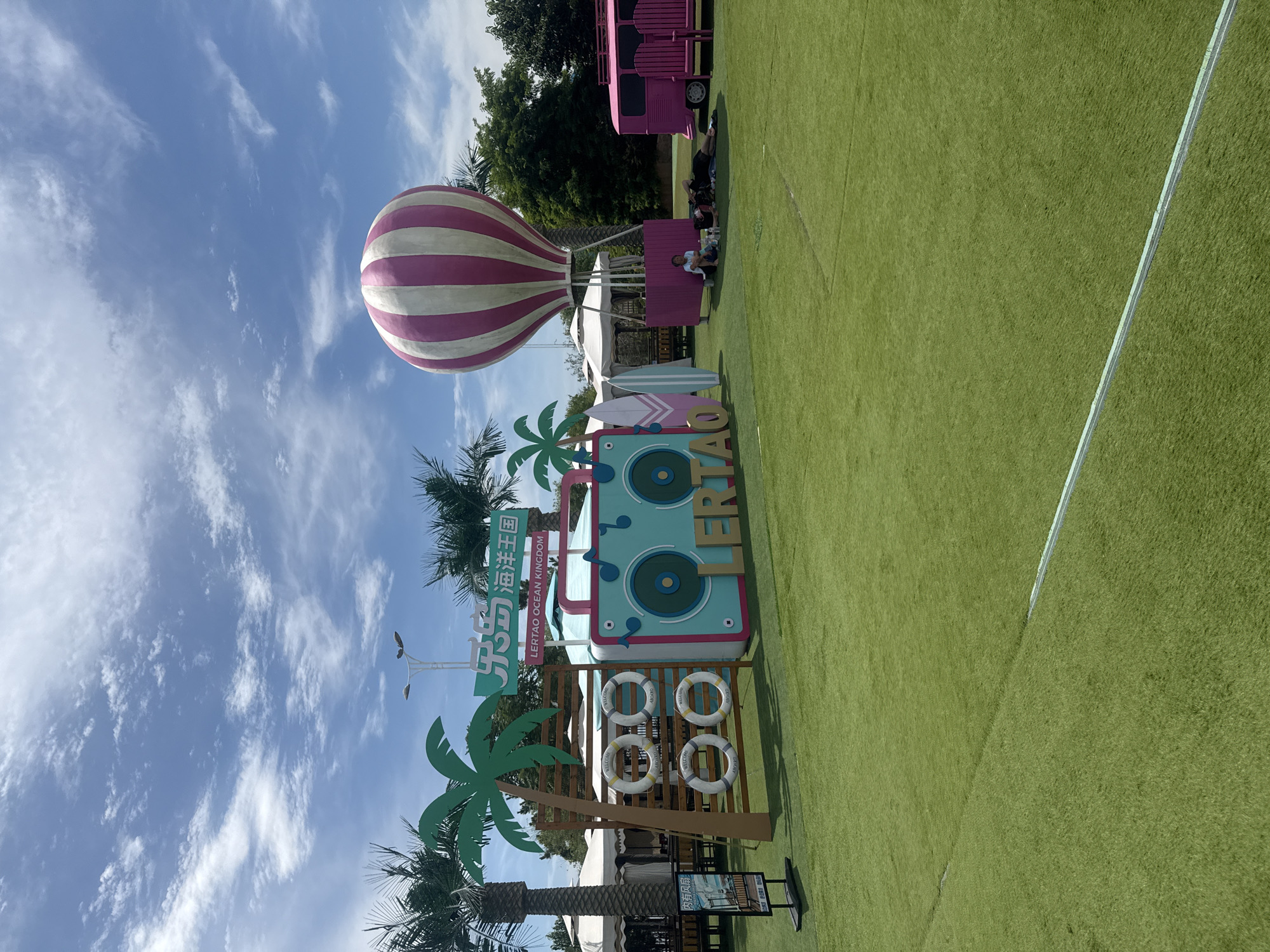 The image depicts a sunny outdoor scene with a bright blue sky filled with scattered white clouds. In the foreground, a meticulously maintained, vibrant green artificial turf covers the ground. To the left, a wooden structure displays four life preservers, each adorned with the words "WELCOME ABOARD" and "WELCOME ABOARD". Palm trees flank this structure, adding to a tropical ambiance. A large, colorful, stylized boombox decoration is prominently featured, with the word "LERTAO" spelled out in large, bold letters below it. Two surfboards stand upright next to the boombox. Further to the right, a large pink and white striped hot air balloon is tethered and rises into the sky. Beneath the balloon, a pink camper van is parked, accompanied by two empty pink chairs. Several people are relaxing on the grass near the van, with one person sitting upright and two others lying down. In the background, white tents and trees complete the scene. A sign with Chinese characters and the English text "LERTAO OCEAN KINGDOM" is visible above the boombox decoration, suggesting the location is Lertao Ocean Kingdom in Qinhuangdao, China.