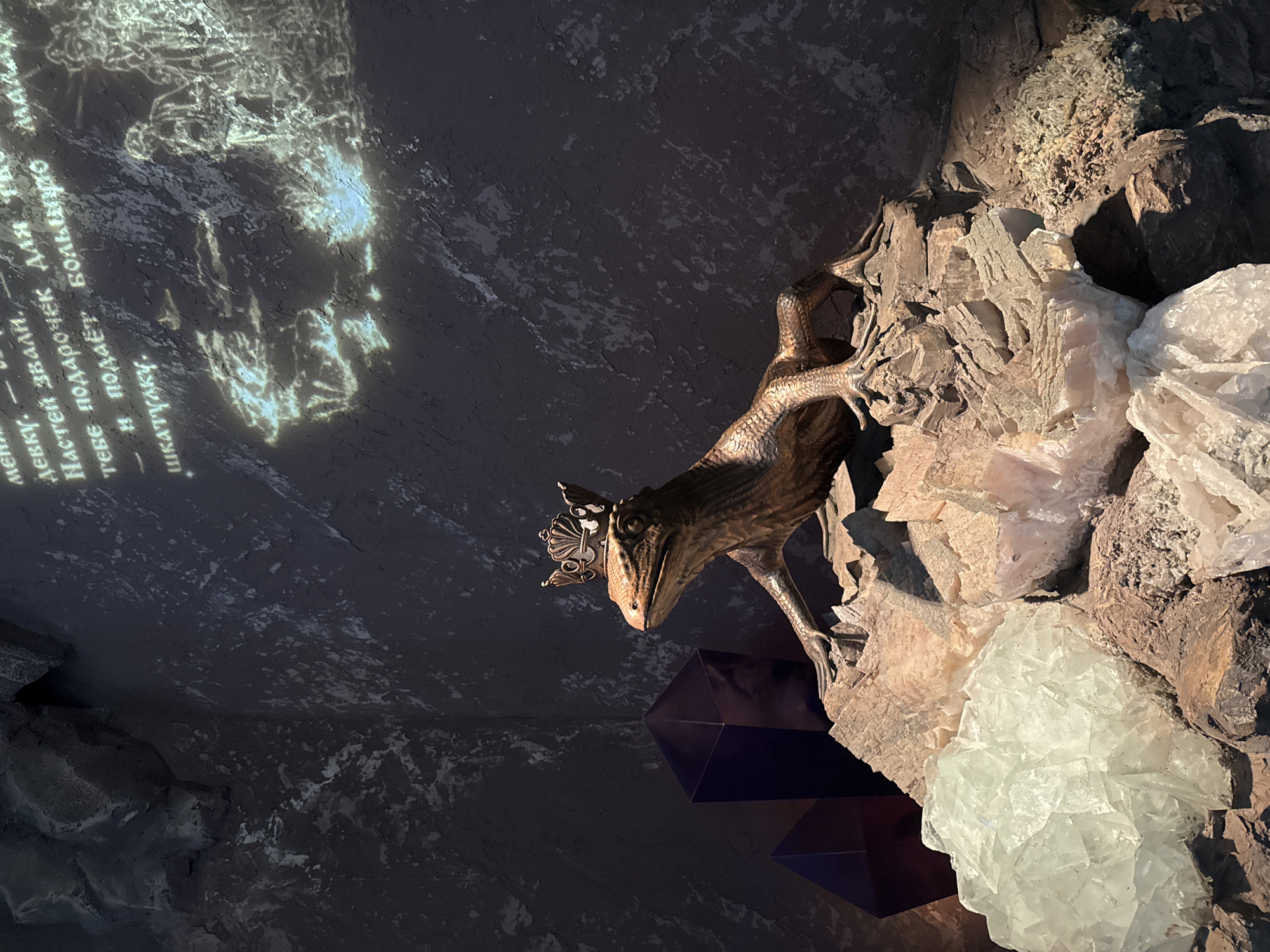 This image captures a bronze sculpture of a lizard wearing a crown, perched atop a rocky outcrop adorned with large crystals. The sculpture is positioned in what appears to be a museum or exhibition space, as indicated by the projected text and imagery on the dark, textured wall behind it. The text, in Russian, is partially legible and seems to be a narrative or poetic excerpt. To the left of the lizard sculpture, two large, dark purple crystal formations are visible. The crystals at the base of the sculpture are a mix of white and pale pink, with some appearing translucent. The overall lighting is dim, with spotlights focused on the sculpture and the crystals, creating dramatic shadows and highlights. The scene is situated in the Museum of Russian Underground Art in Moscow, Russia.