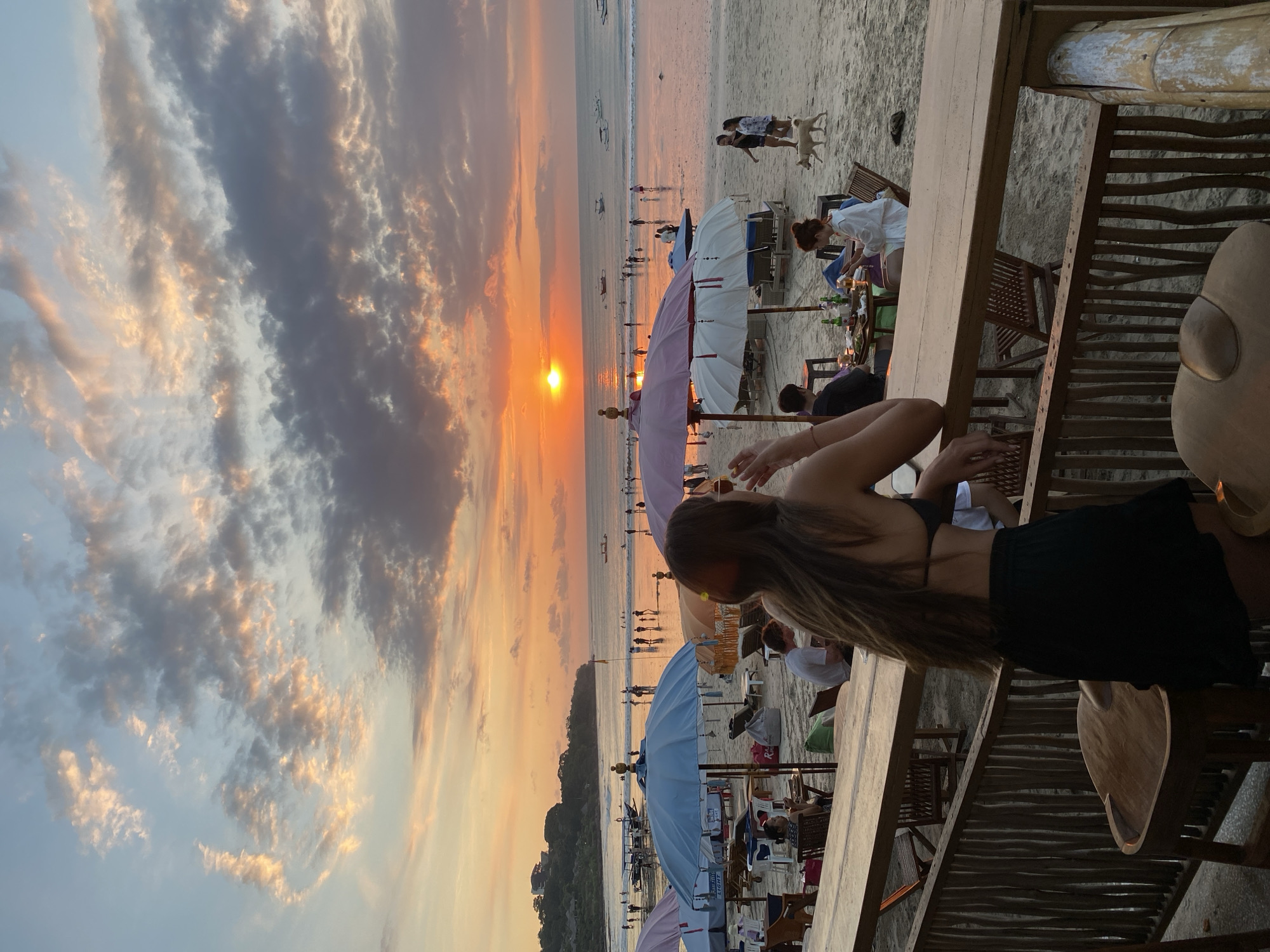 The image presents a vibrant sunset scene at a bustling beach, viewed from a raised wooden patio or restaurant deck. The sun, a brilliant orange sphere, is low on the horizon, casting a warm glow across the sky and ocean. The sky features dramatic clouds, transitioning from fiery oranges and yellows near the sun to softer pinks, purples, and deep greys higher up.
In the foreground, the back of a person with long brown hair, wearing a black top and skirt, is visible, leaning on a wooden railing. This person is observing the activity below. Other patrons are seated at wooden tables on the patio, some with bottles of drinks, enjoying the evening.
The sandy beach below is active with numerous people. Several large beach umbrellas in shades of white, light blue, and pinkish-purple are set up, shading groups relaxing on lounge chairs. Many individuals are seen strolling along the shoreline or wading in the shallow waters. In the mid-ground, a woman in a white shirt attends to a table, and two dogs walk on the sand nearby. Small boats are visible in the ocean, and a distant hilly landmass can be seen on the left horizon.
The setting evokes a relaxed and leisurely atmosphere, characteristic of a tropical beach destination like Kuta Selatan, Indonesia, during the golden hour. On a light blue beach umbrella, the text "Bin-tang LIGHT" is faintly visible. The scene captures the beauty and enjoyment of a sunset beach experience.