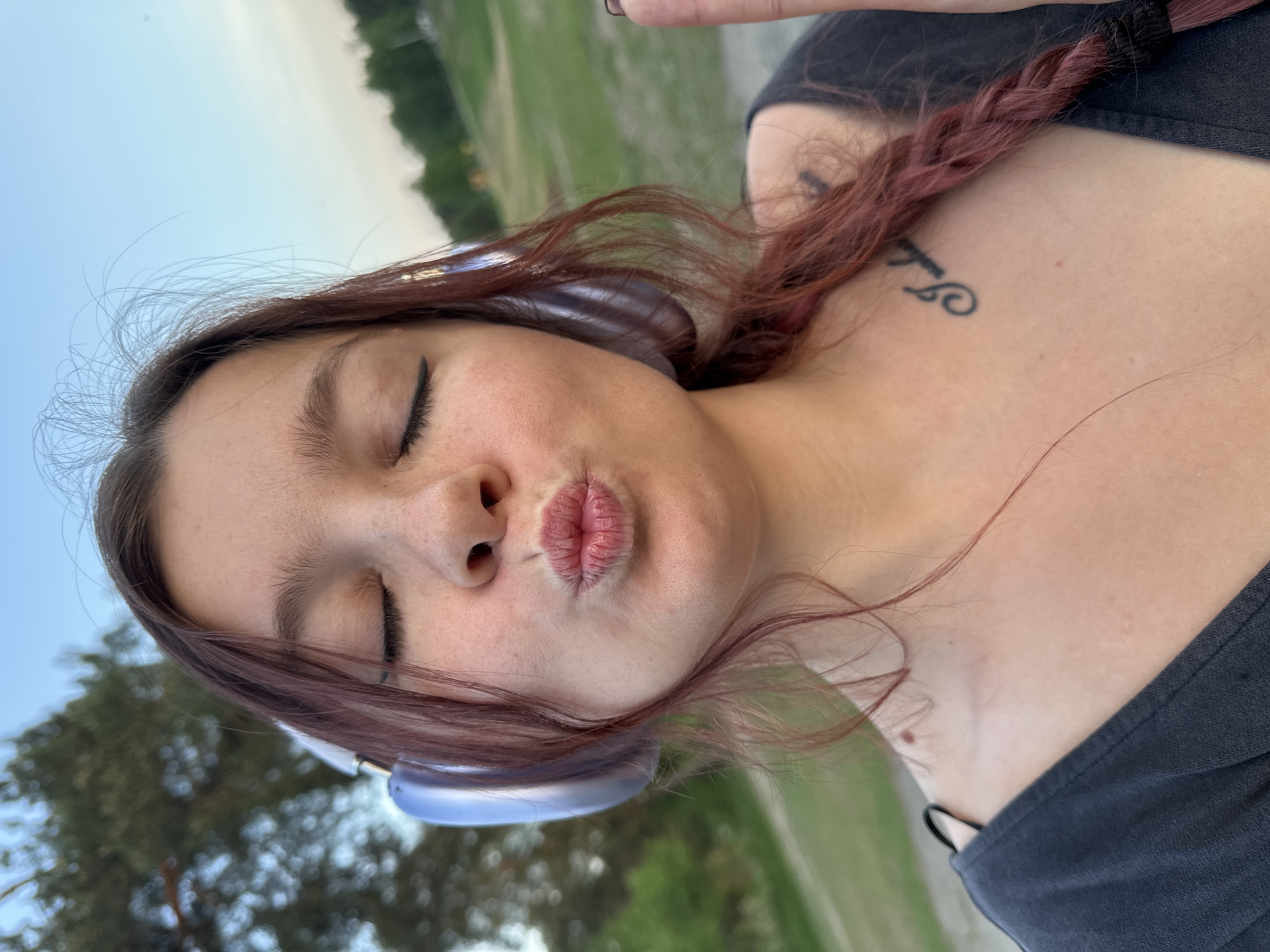 A young woman is posing outdoors. She has light skin, dark eyebrows, and closed eyes. Her lips are pursed in a kissing expression. She is wearing headphones and a dark-colored top. Her hair is dyed red and partially braided. A tattoo is visible on her upper chest near her shoulder. The background appears to be a park or grassy area with trees. The location is not Vladimir, Russia, because no location is mentioned in the image.