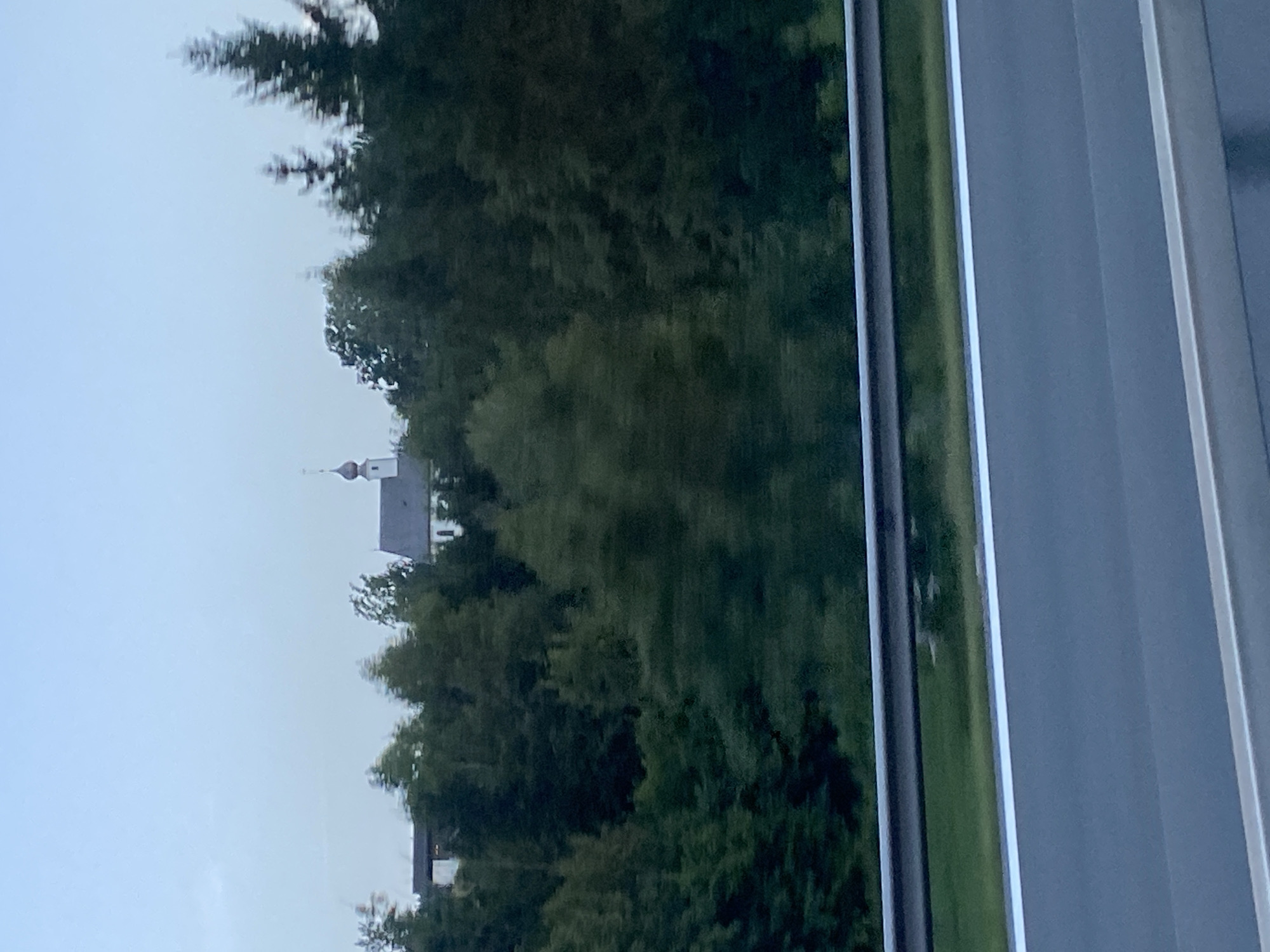 The image shows a blurred view of a church with a domed roof and a cross, situated behind a dense line of dark green trees. To the left of the church, a building with a light-colored facade is partially visible. In the foreground, a highway guardrail runs horizontally across the lower portion of the image, with a grey asphalt road and white lane markings visible below it. The sky above is a pale, overcast blue. The image appears to have been taken from a moving vehicle, as indicated by the motion blur. The scene is located in or near Siegsdorf, Germany.