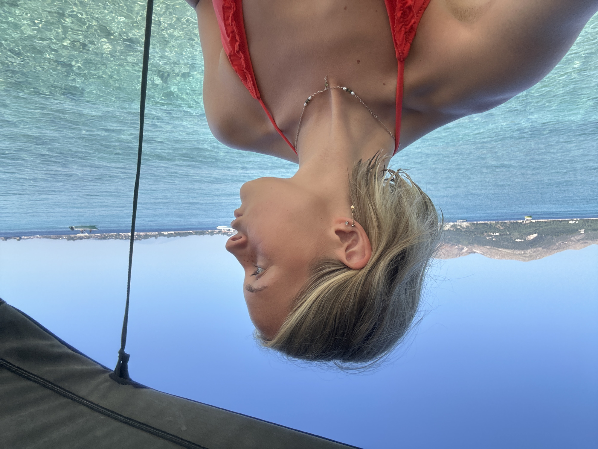 A young woman with short blonde hair and fair skin, wearing a red bikini and a silver necklace, looks to the right. Her face is partially obscured by her hair. She is on a boat with a dark canopy overhead. The background shows clear turquoise water, a distant hilly coastline, and a bright blue sky. A small white boat is visible in the distance on the water. The location appears to be the coastal area of Laganas Municipal Unit, Greece.