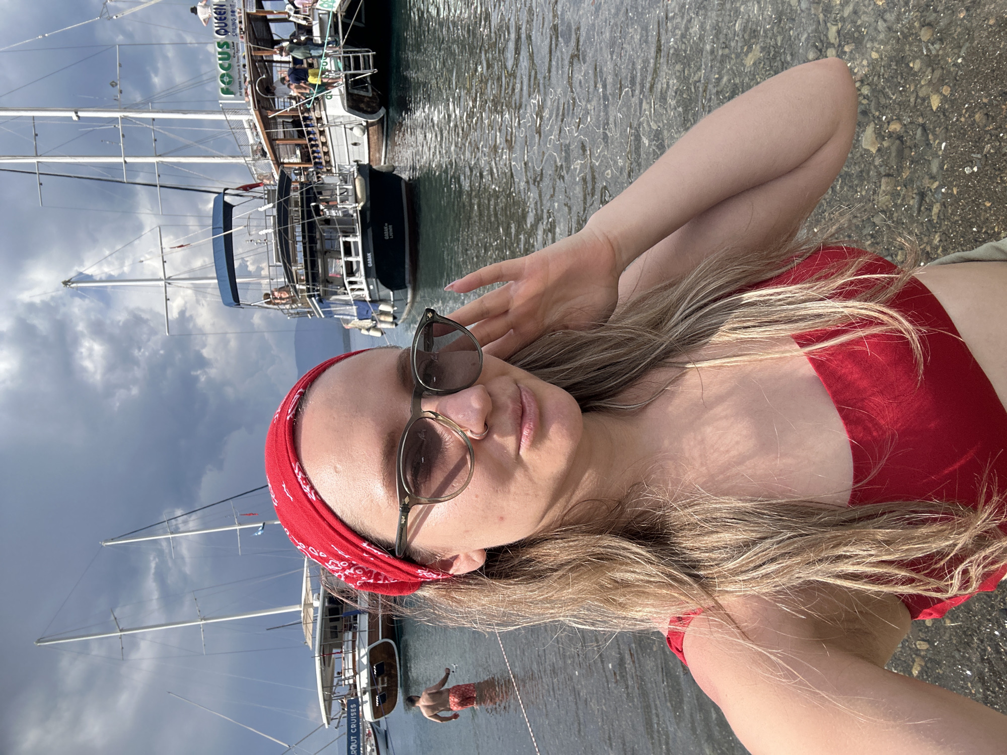 The image captures a woman with long blonde hair, wearing sunglasses and a red bandana, posing in front of a harbor setting in Fethiye, Turkey. She is wearing a red top, possibly a swimsuit. Several tour boats are docked in the background, displaying names such as "Focus Queen" and "Garot Cruises." The water is clear and calm, and a man is seen standing in the shallow water. The beach is pebbly. The sky is overcast.