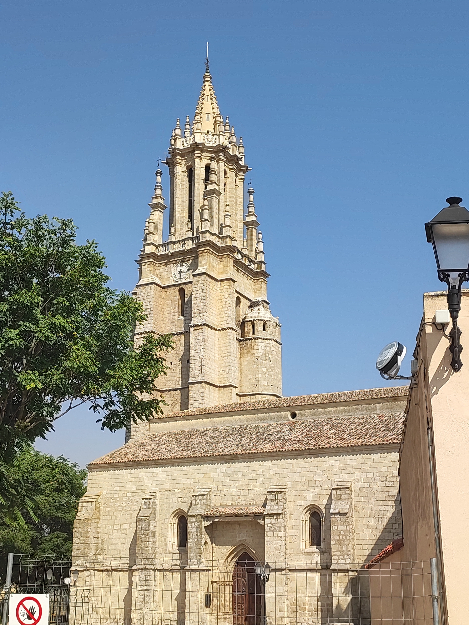 The image shows a historic church with a tall bell tower in Ampudia, Spain. The church is constructed of light-colored stone and features a red-tiled roof. The bell tower is intricately designed with spires and decorative elements, and a clock face is visible on one of its sides. To the left of the tower, a lush green tree partially obscures the view. On the right side of the image, a section of a light-colored building with a street lamp is visible. A metal fence runs along the bottom of the image, and a white sign with a red circle and a diagonal line, indicating a prohibition, is present on the left side of the fence. The sky is clear and blue.