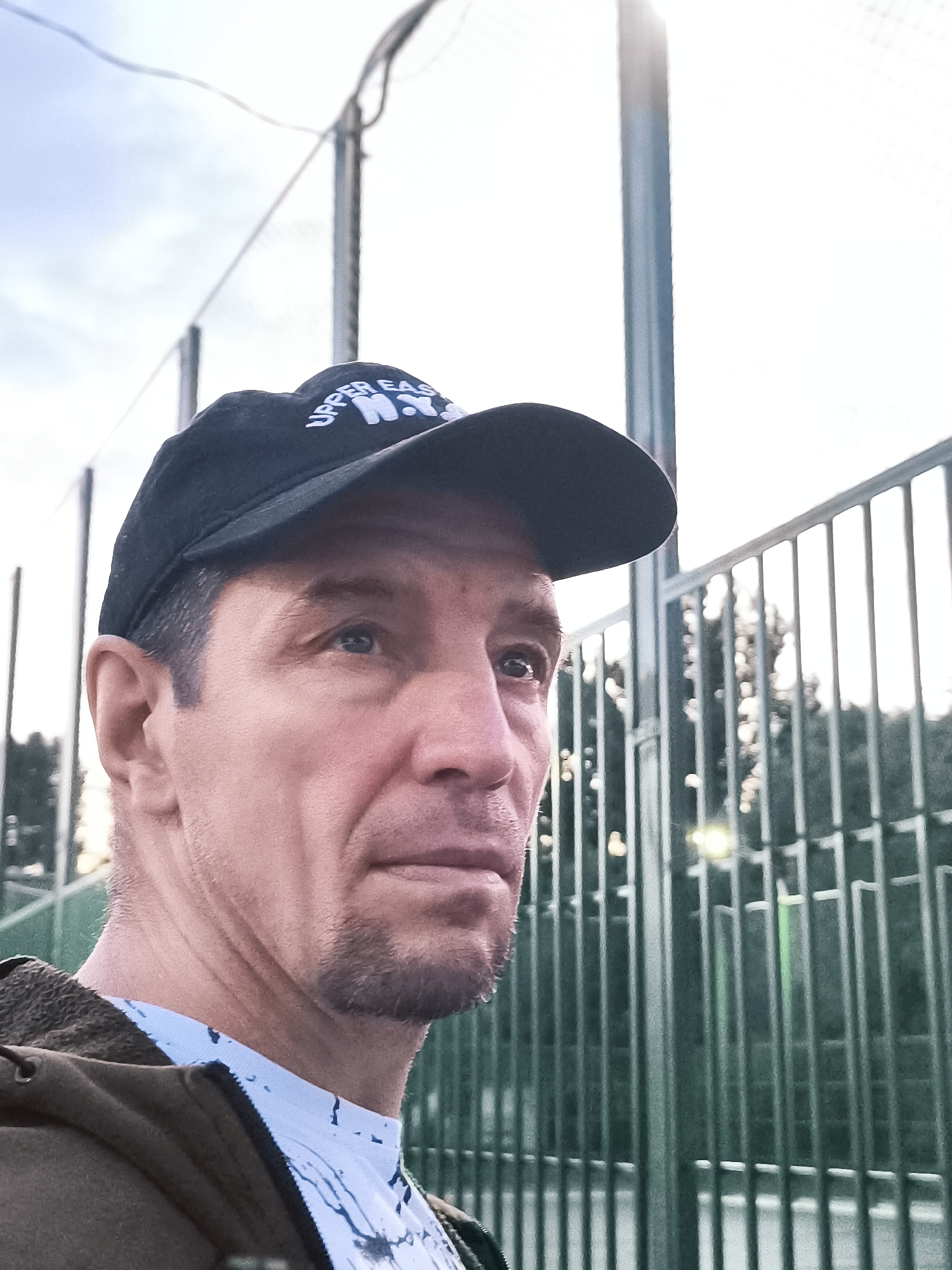 A medium shot photo, taken from the chest up, shows a man wearing a black baseball cap with white lettering that reads "UPPER EAST N.Y.". The man is looking to his right, with his head slightly tilted up. His skin is a light tone, and he has short, dark hair. He is wearing a light blue shirt with a dark pattern, and a brown jacket is visible over his left shoulder. The background consists of a green metal fence, with trees and a bright sky visible beyond. The lighting is soft, suggesting it may be evening or early morning. The image appears to be taken outdoors, possibly in a park or recreational area. There is no indication of the city of Qarağandı, Kazakhstan, or its city administration in this image.