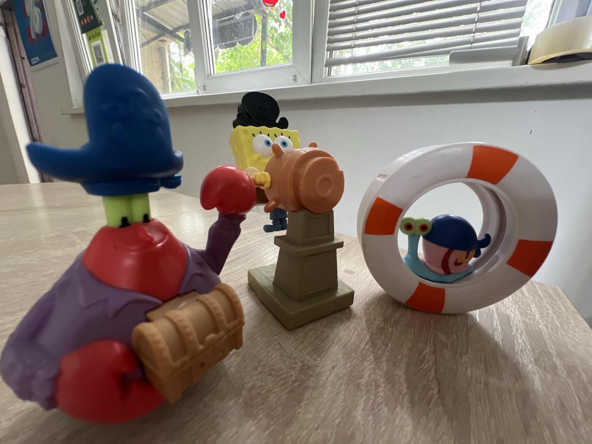 The image displays a collection of Spongebob Squarepants themed toys arranged on a wooden surface, likely a table. In the foreground to the left is a toy resembling Mr. Krabs, distinguished by its red claws, purple shirt, and a blue pirate-like hat. It is holding a small, wooden treasure chest. To the right of Mr. Krabs, a Spongebob Squarepants toy is depicted wearing a black hat and holding a brown steering wheel-like object. Behind Spongebob is a white life preserver with orange stripes, through which a toy resembling Gary the Snail is visible, wearing a blue cap. The background consists of a window with blinds, and an outdoor view of trees and possibly some buildings. The lighting suggests an indoor setting during the daytime. The specific location in Almaty, Kazakhstan cannot be determined from the image.