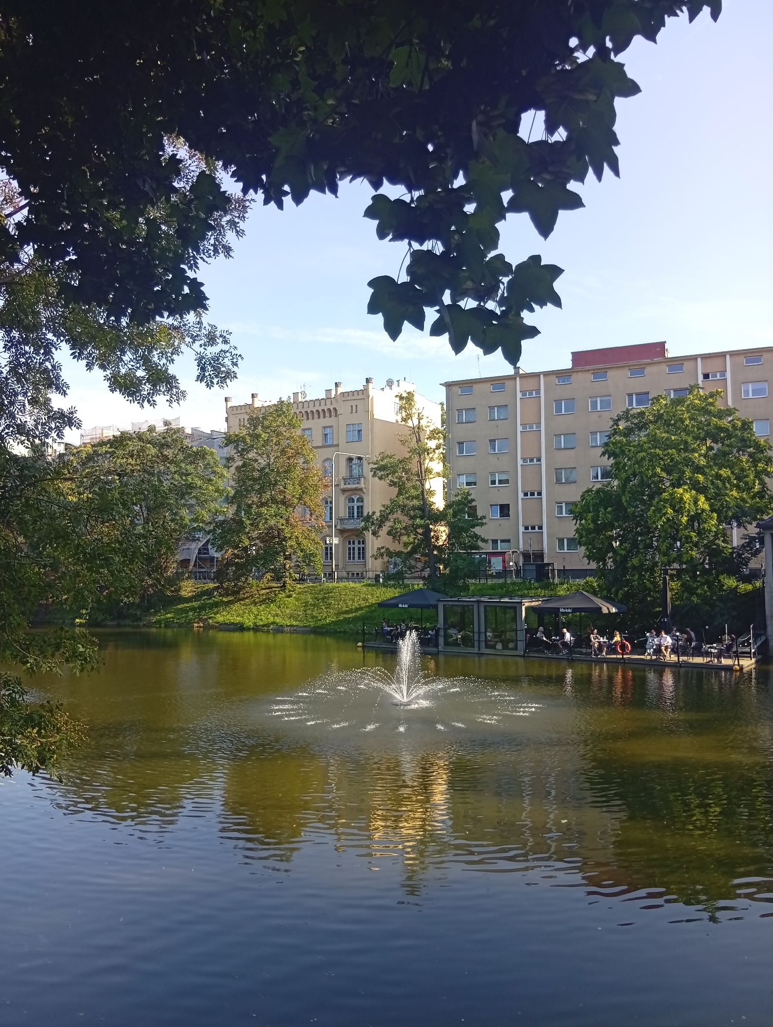 A pond with a fountain is in the foreground. A pier with people sitting at tables and umbrellas is on the right side of the pond. Behind the pond, there are trees and buildings. One building is light yellow with a red roof and ornate windows and balconies. Another building behind it is taller and grey with many windows. The scene is set in Wrocław, Poland, specifically in the area of Partynice, near the Wrocław Partynice Racecourse, where a waterside relaxation zone has been developed.